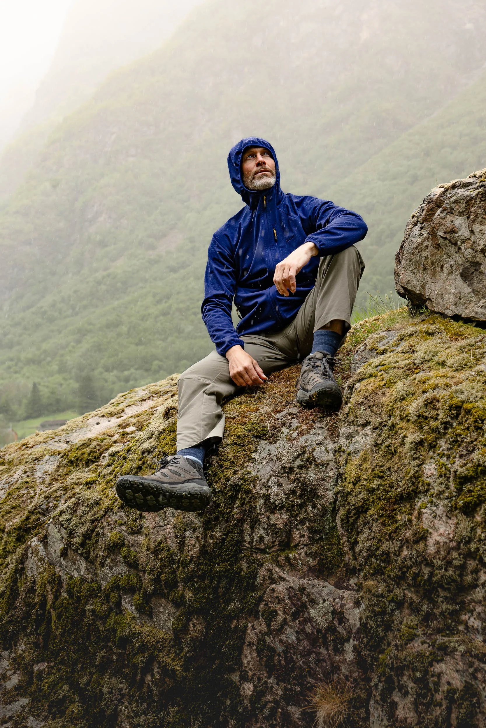 A man dressed in outdoor gear with a blue rain jacket and hiking boots is sitting on a moss-covered rock on a mountain slope, surrounded by fog and lush greenery.