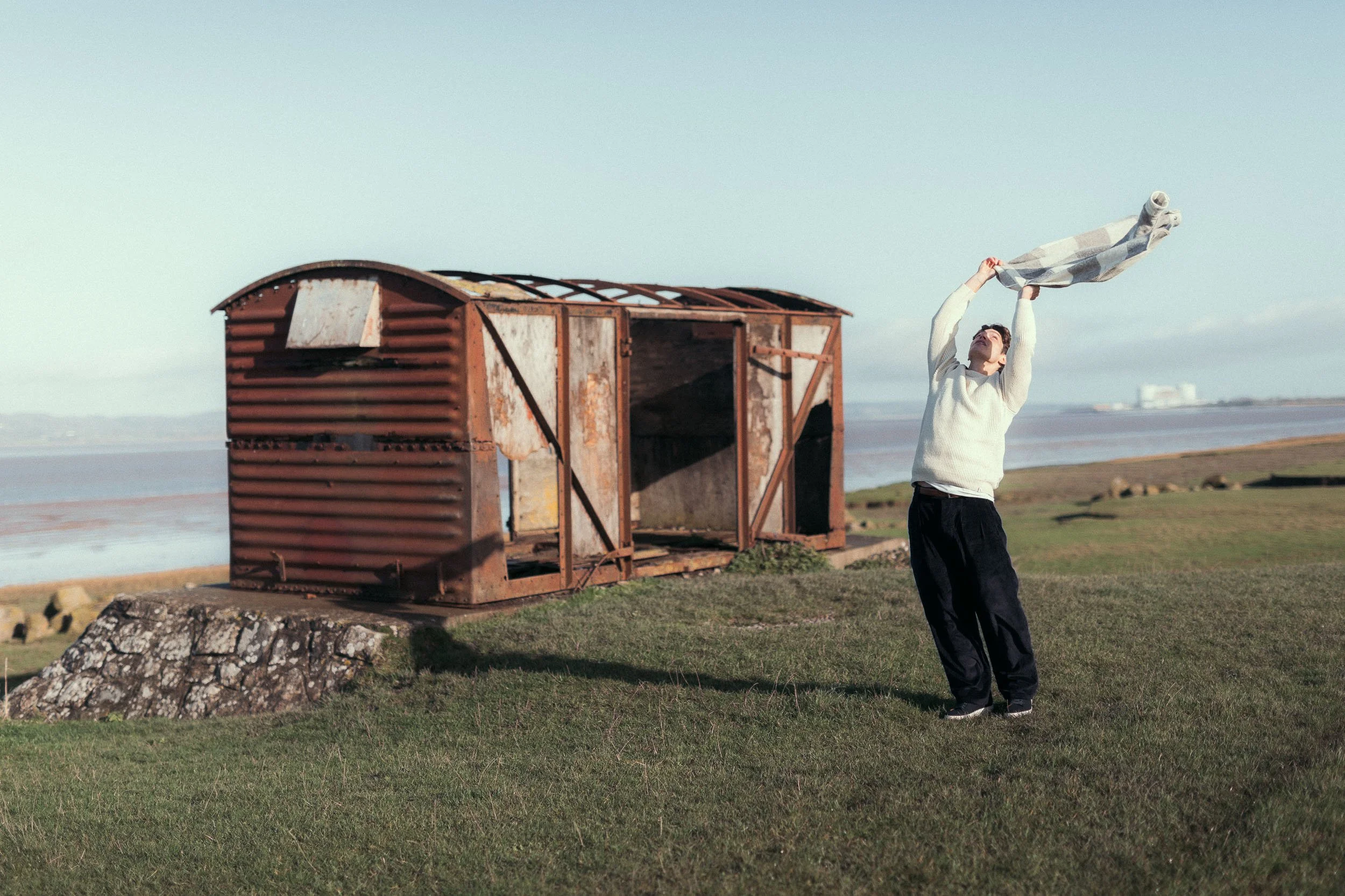 A young man is outside near the coast, standing on green grass and holding a checkered fabric in the air. There is a rusty, weathered small wooden shack behind him and a large rock to the left. The ocean and some buildings are visible in the background, under a clear sky.