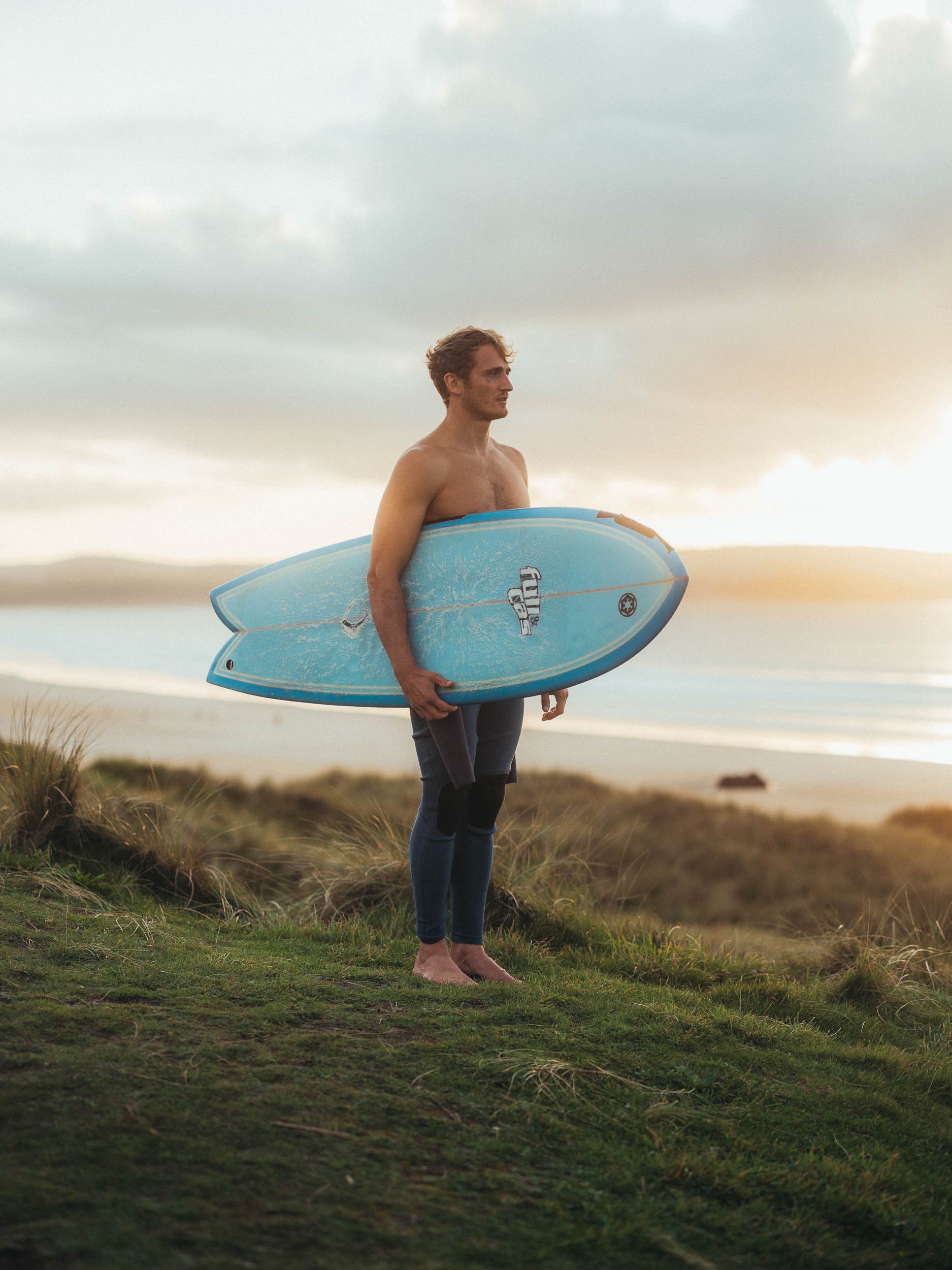 A man standing barefoot on grass, holding a blue surfboard under his right arm, with ocean and cloudy sky in the background.