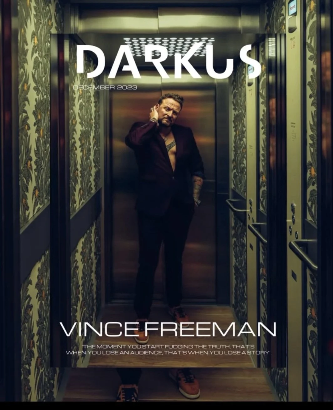 Cover of DARKUS magazine featuring Vince Freeman in an elevator, wearing a black blazer and orange sneakers, standing with one hand on his neck, with patterned wallpaper on the elevator walls.