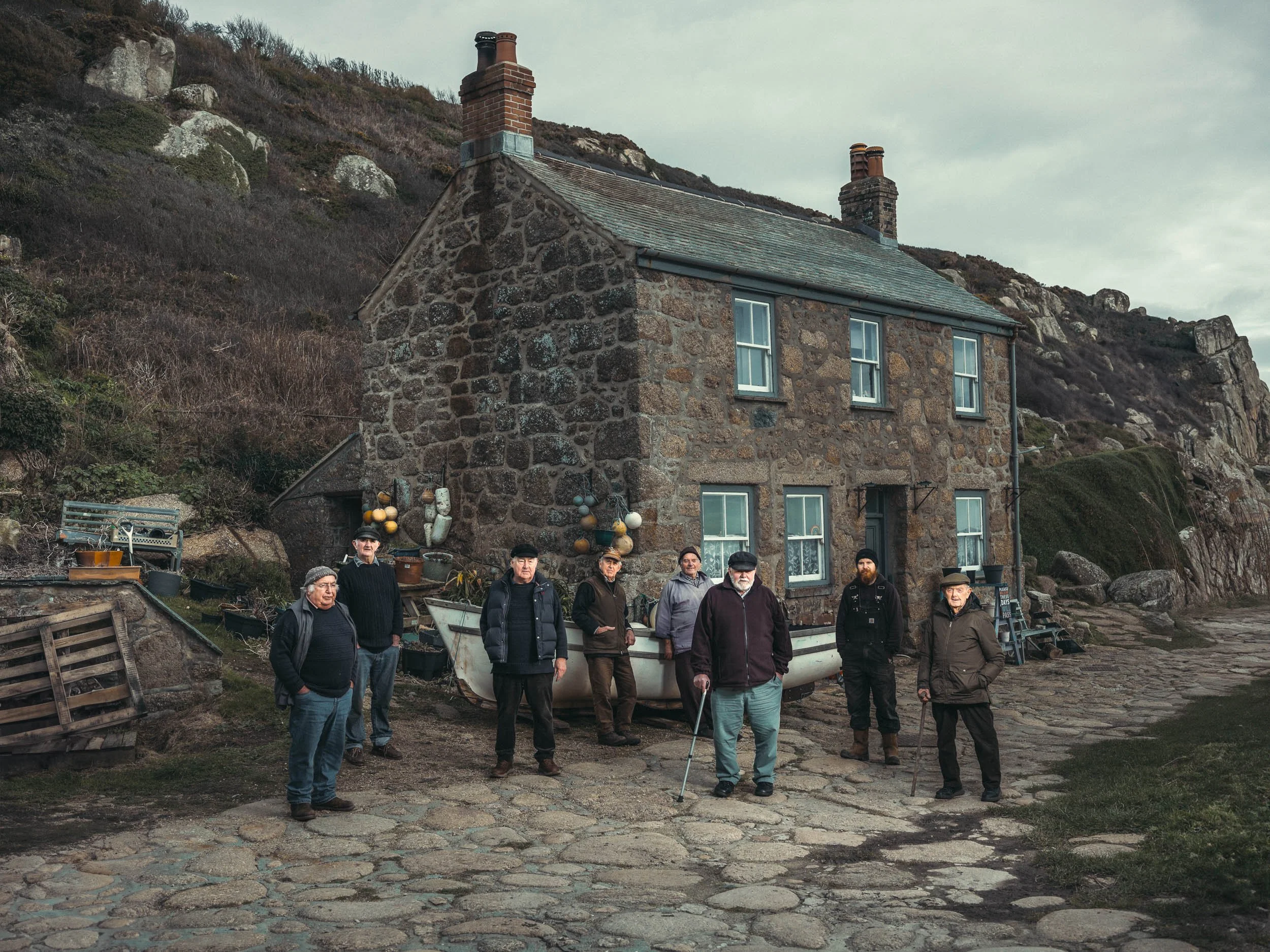 A group of men standing on a stone-paved area outdoors in front of a stone house with three chimneys, behind a rocky hillside, under an overcast sky.
