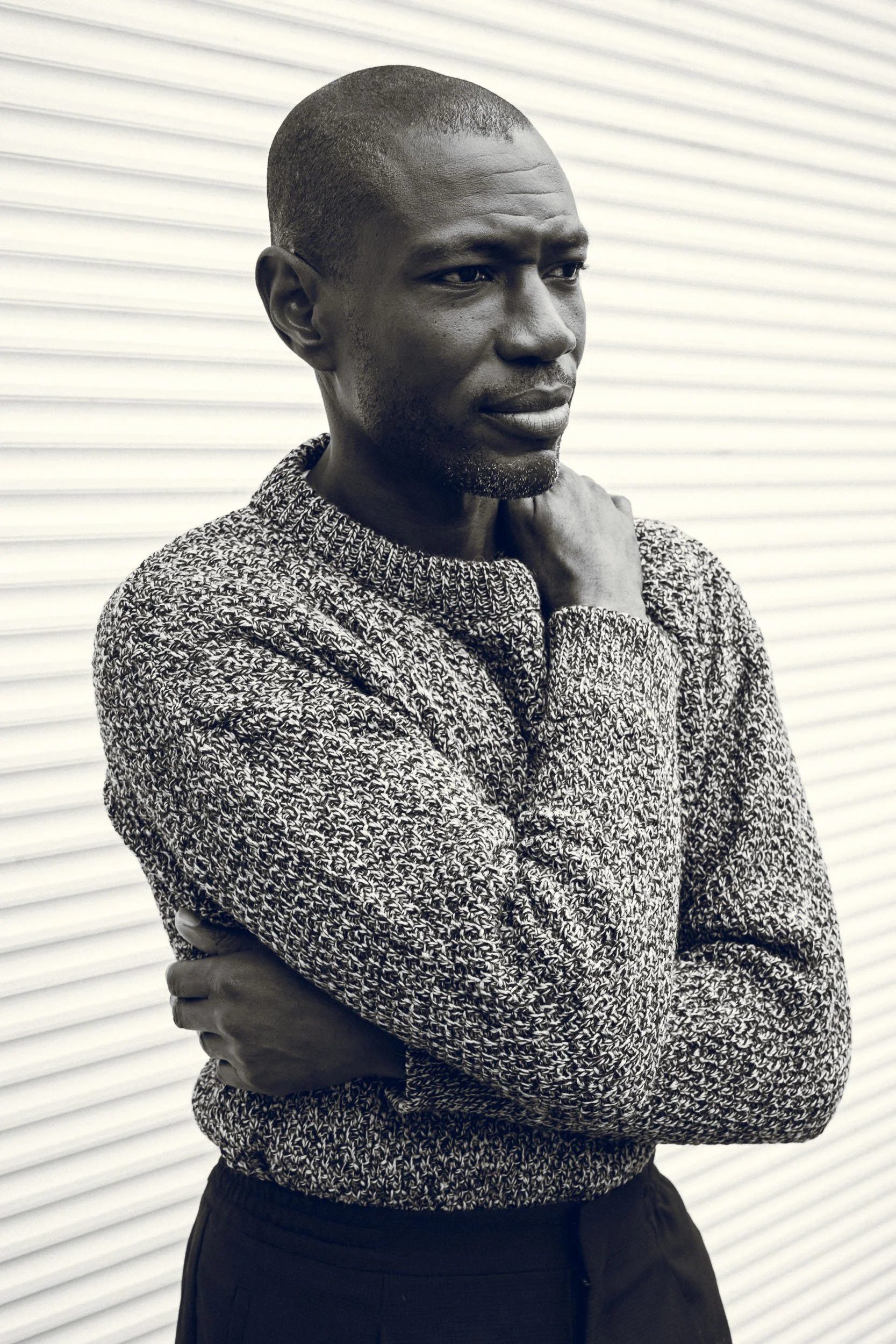A man with dark skin, a shaved head, and a short beard, is wearing a patterned sweater and black pants, standing with his arms crossed in front of a white, corrugated background.