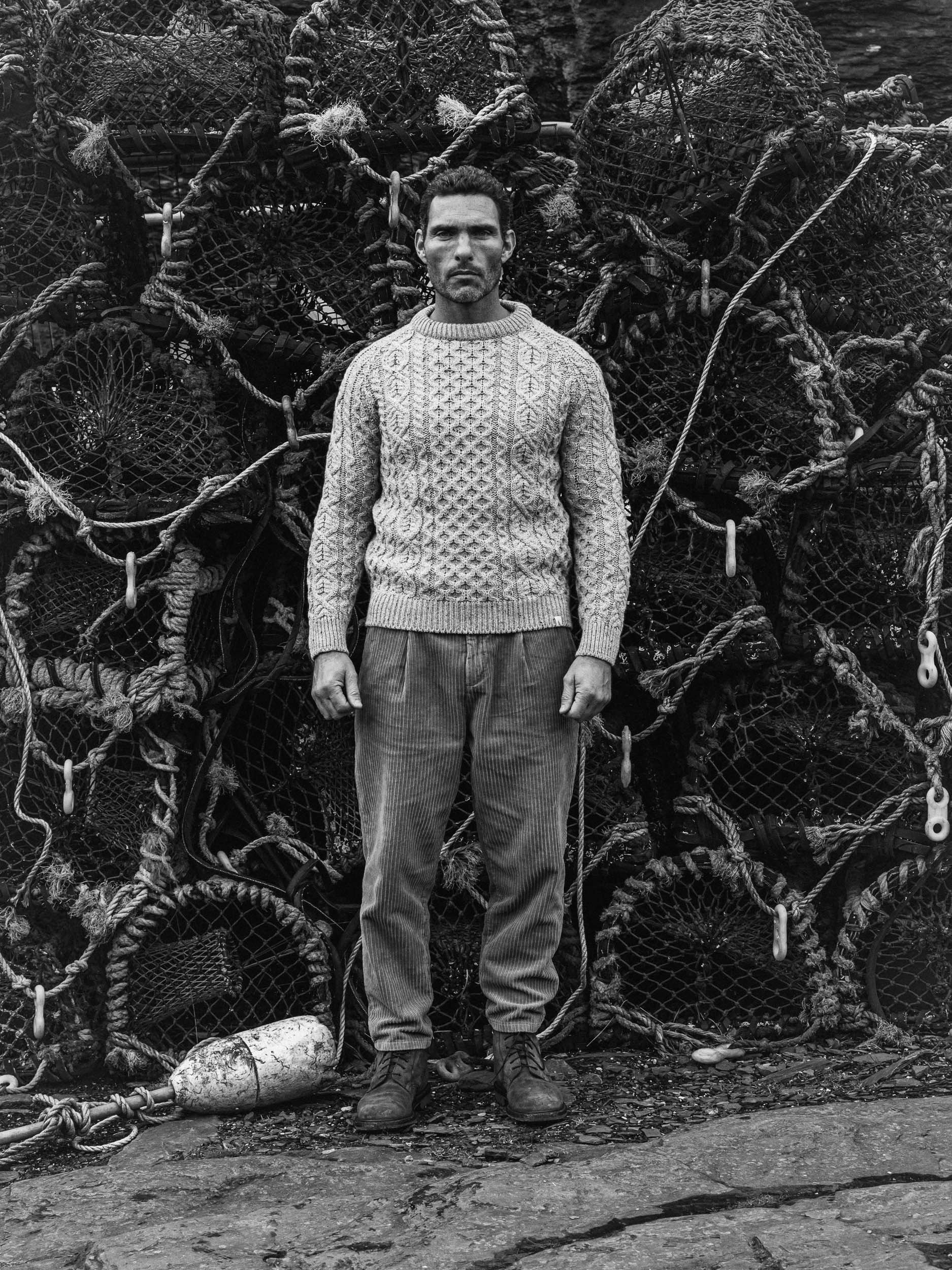 A man standing in front of a pile of lobster traps, wearing a knit sweater, pants, and boots, with a serious expression.