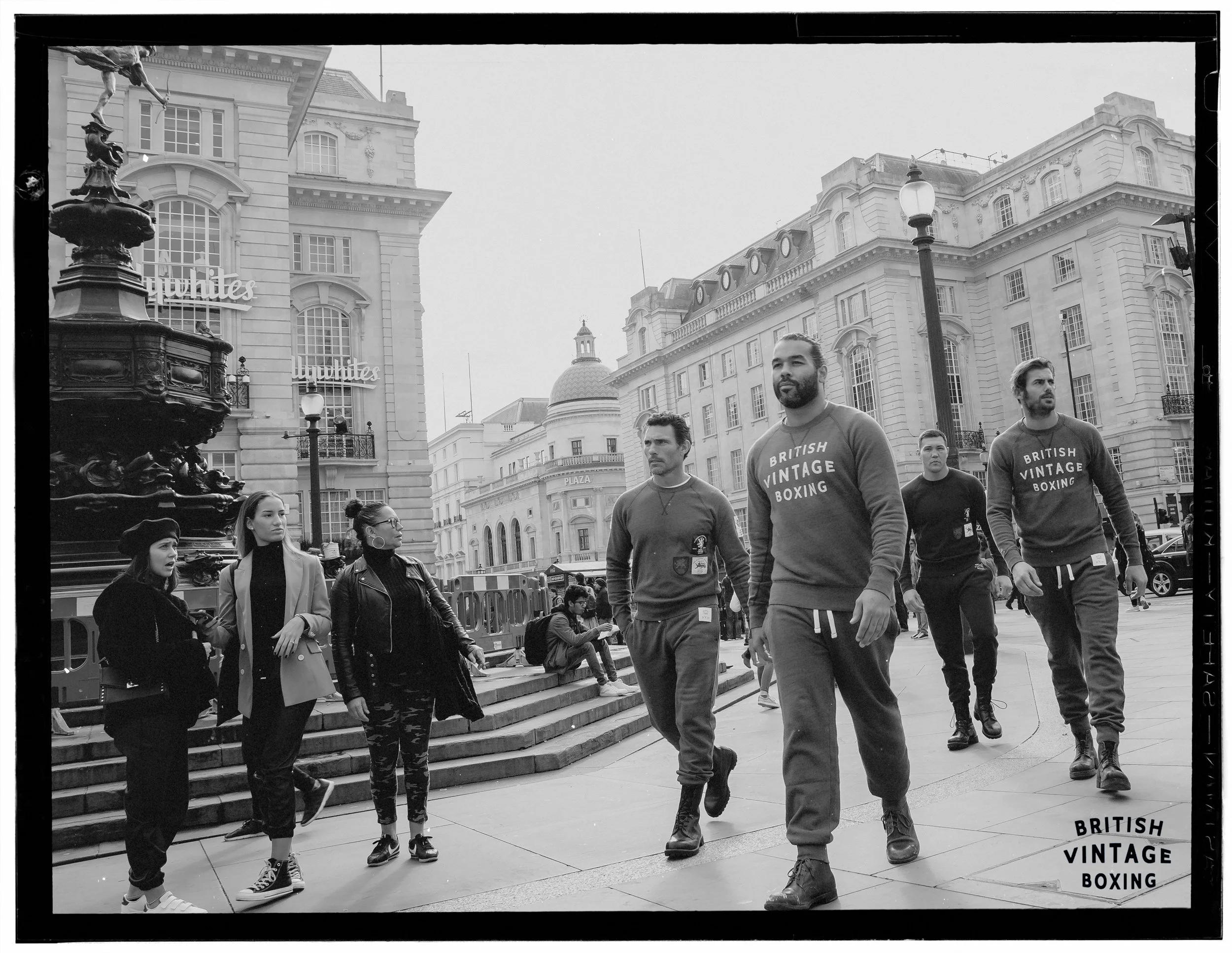 A black and white photo of men and women walking on a city street with classic architecture and a fountain. They are wearing shirts that say 'British Vintage Boxing'.