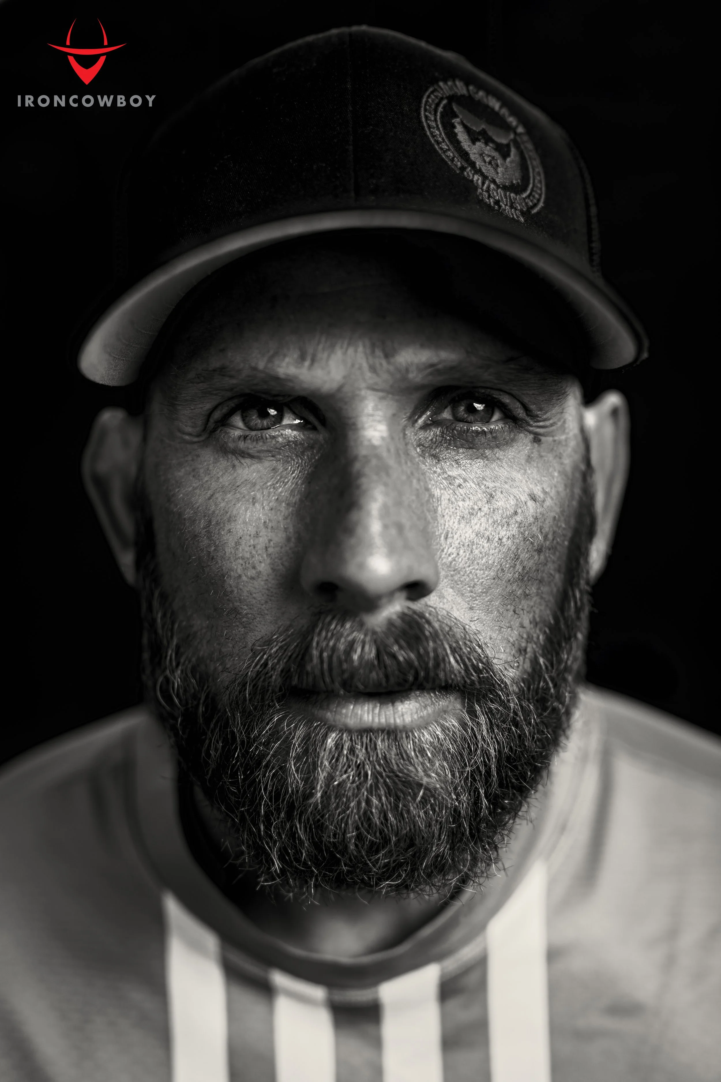 Black and white close-up of a man's face, wearing a baseball cap and hoodie, with intense eyes and a full beard.