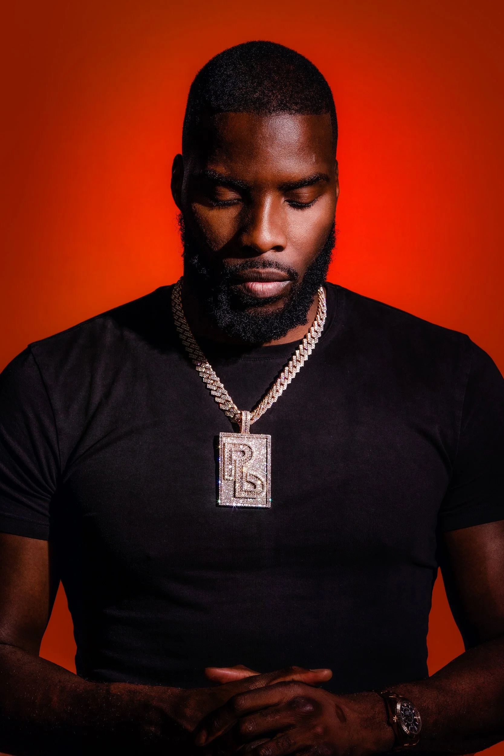 A young man with a short beard and closed eyes, wearing a black t-shirt, a large diamond chain with a pendant shaped like the letter R, and a wristwatch, against a bright orange background.