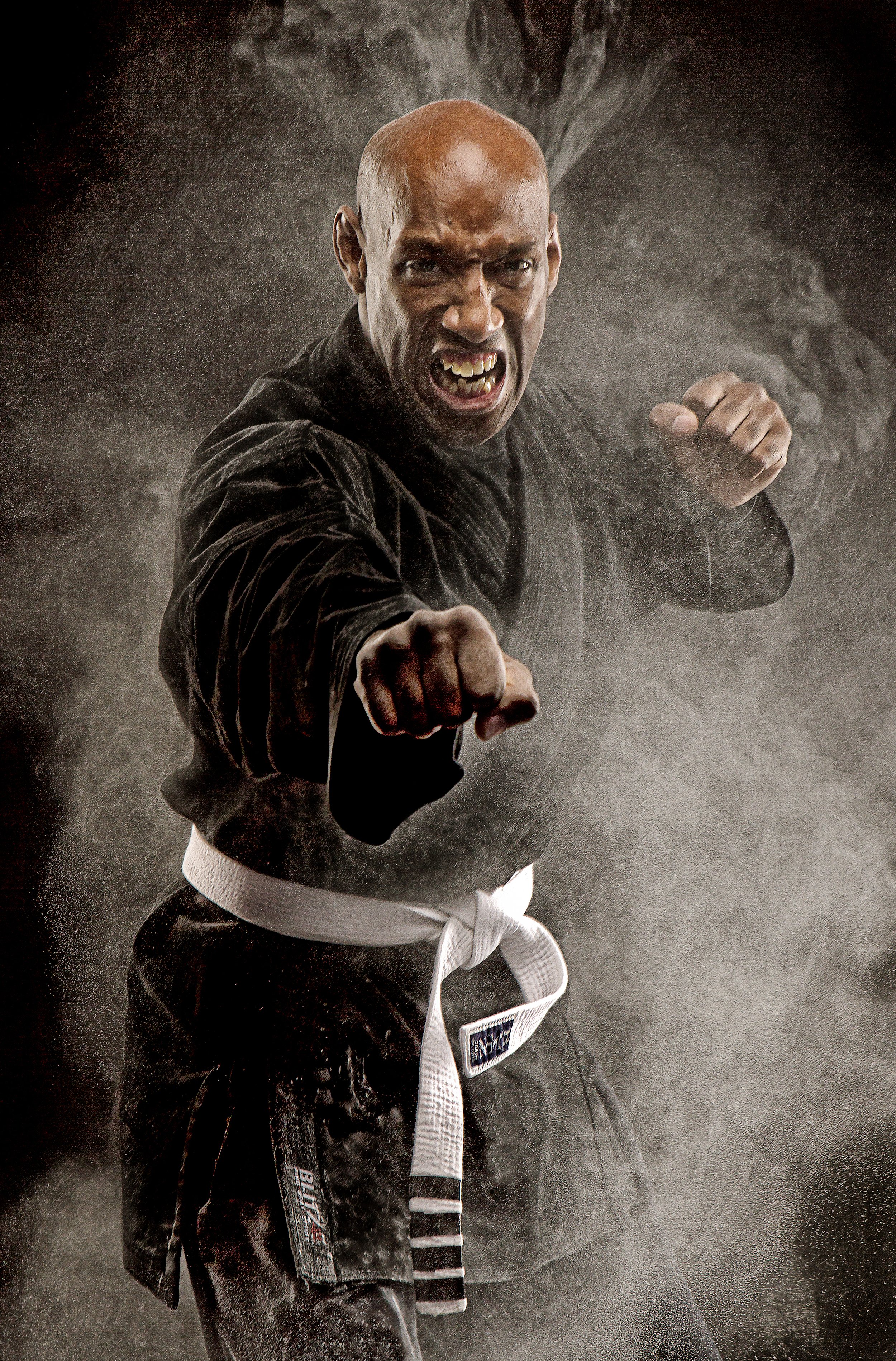 A man in a black martial arts gi with a white belt performs a martial arts move, surrounded by a smoky atmosphere, with an intense facial expression.