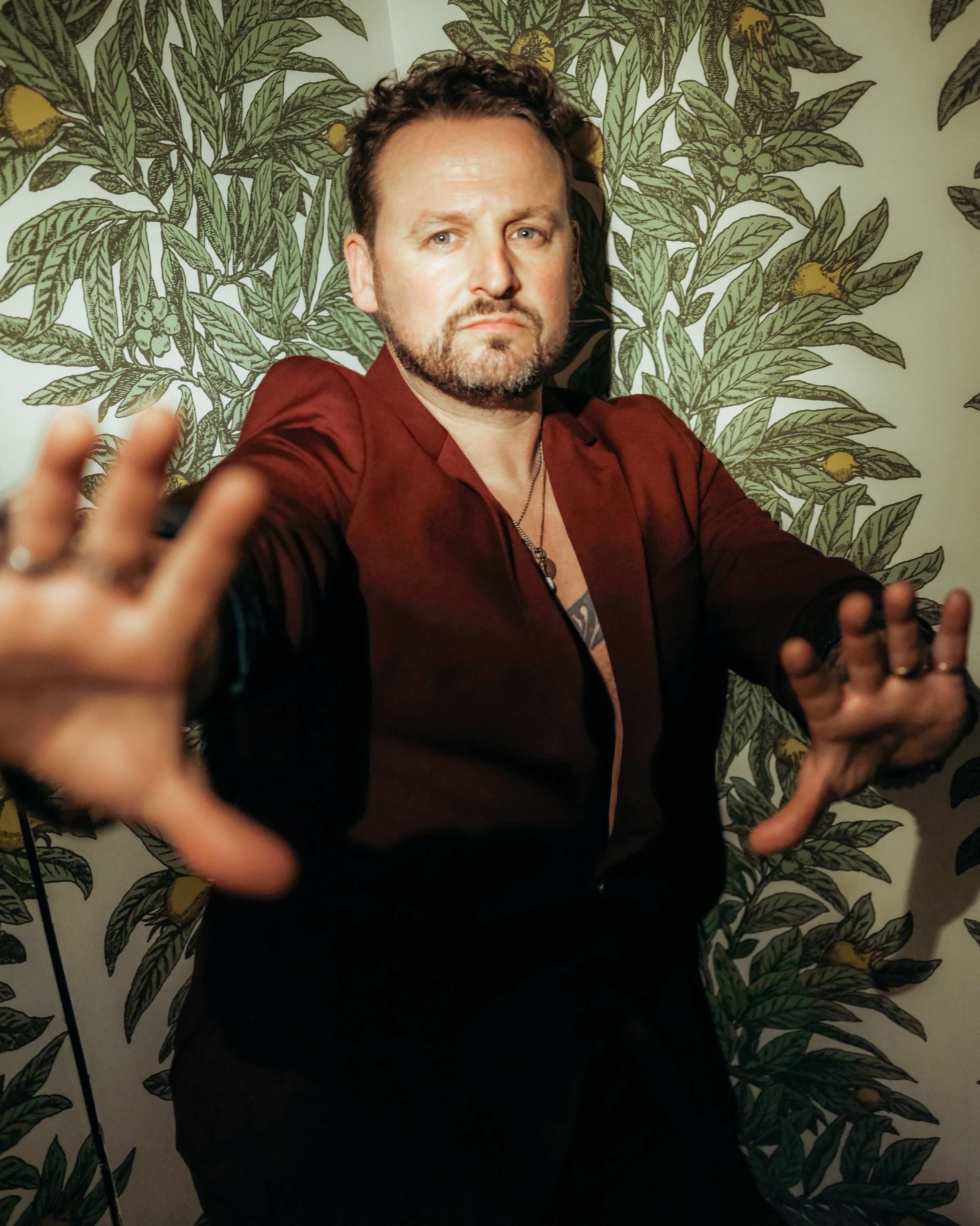 A man with short, dark, curly hair and a beard, wearing a burgundy blazer, taking a selfie in front of a leafy, floral wallpaper. His face is centered, with his hands reaching out towards the camera.