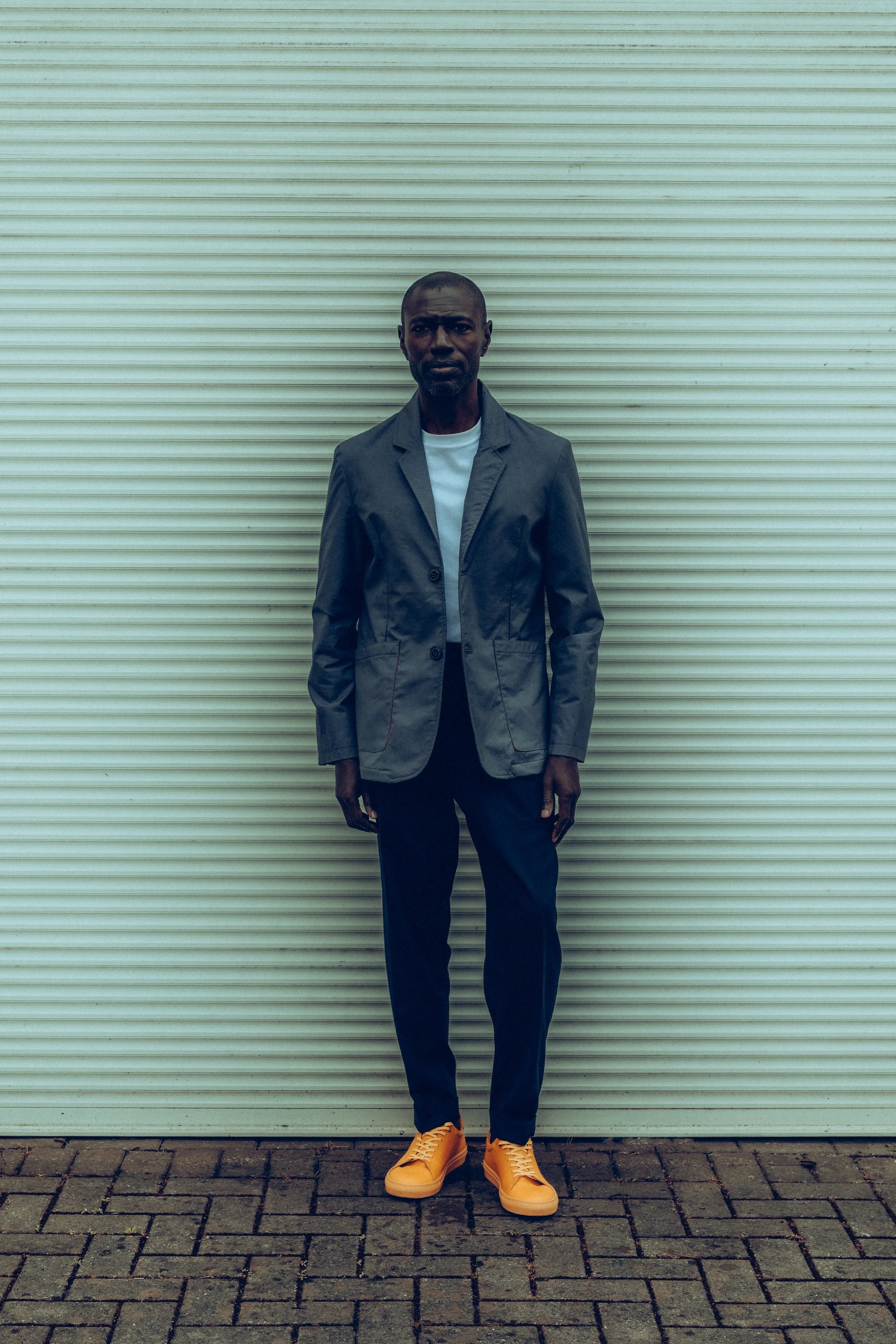 A man standing outdoors in front of a light-colored garage door, wearing a gray blazer, white shirt, dark pants, and yellow sneakers.
