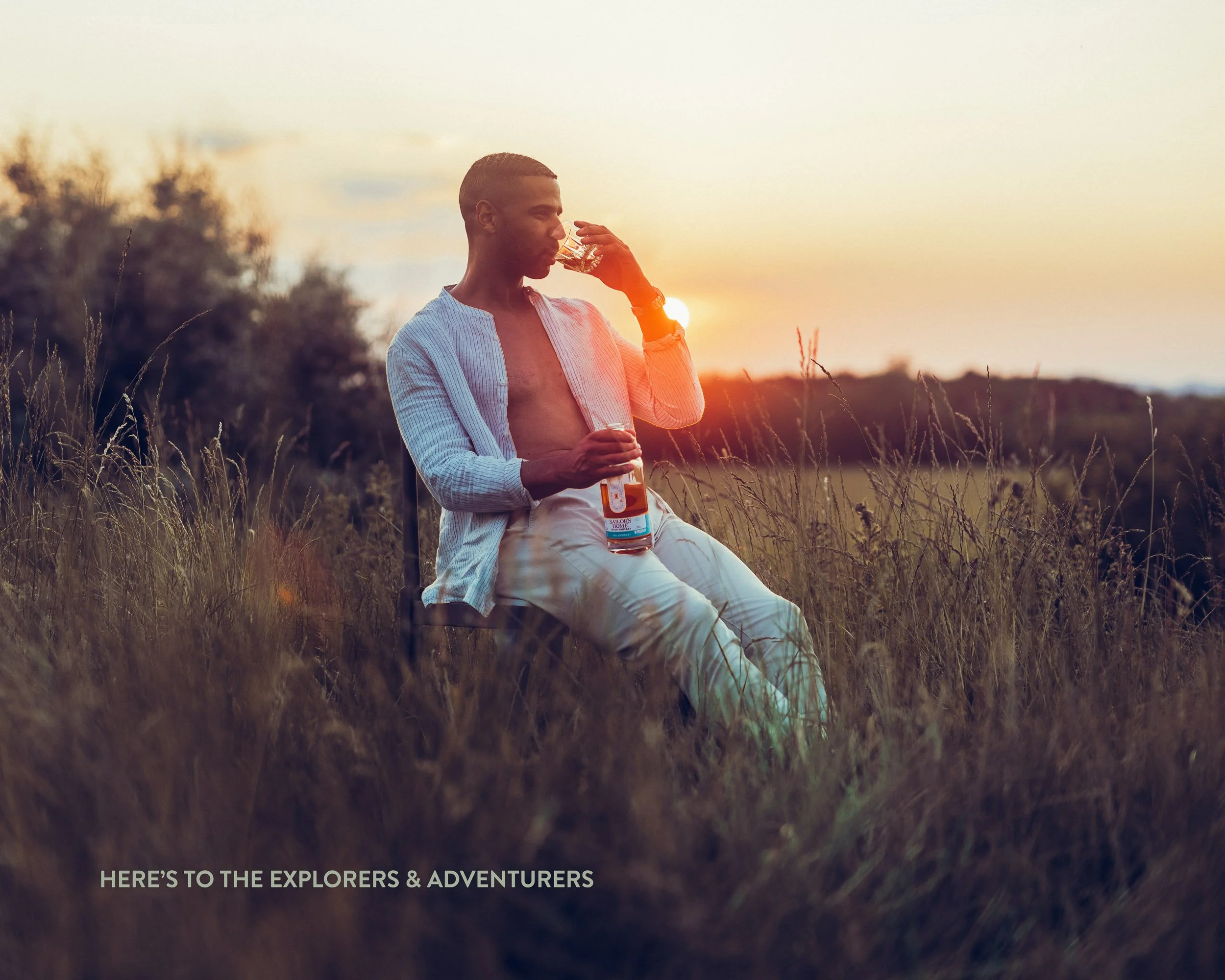 A man sitting in a field at sunset, drinking from a glass, with a partially filled beer bottle resting on his lap. He wears a partially unbuttoned shirt and white pants, with trees and a sunset in the background.