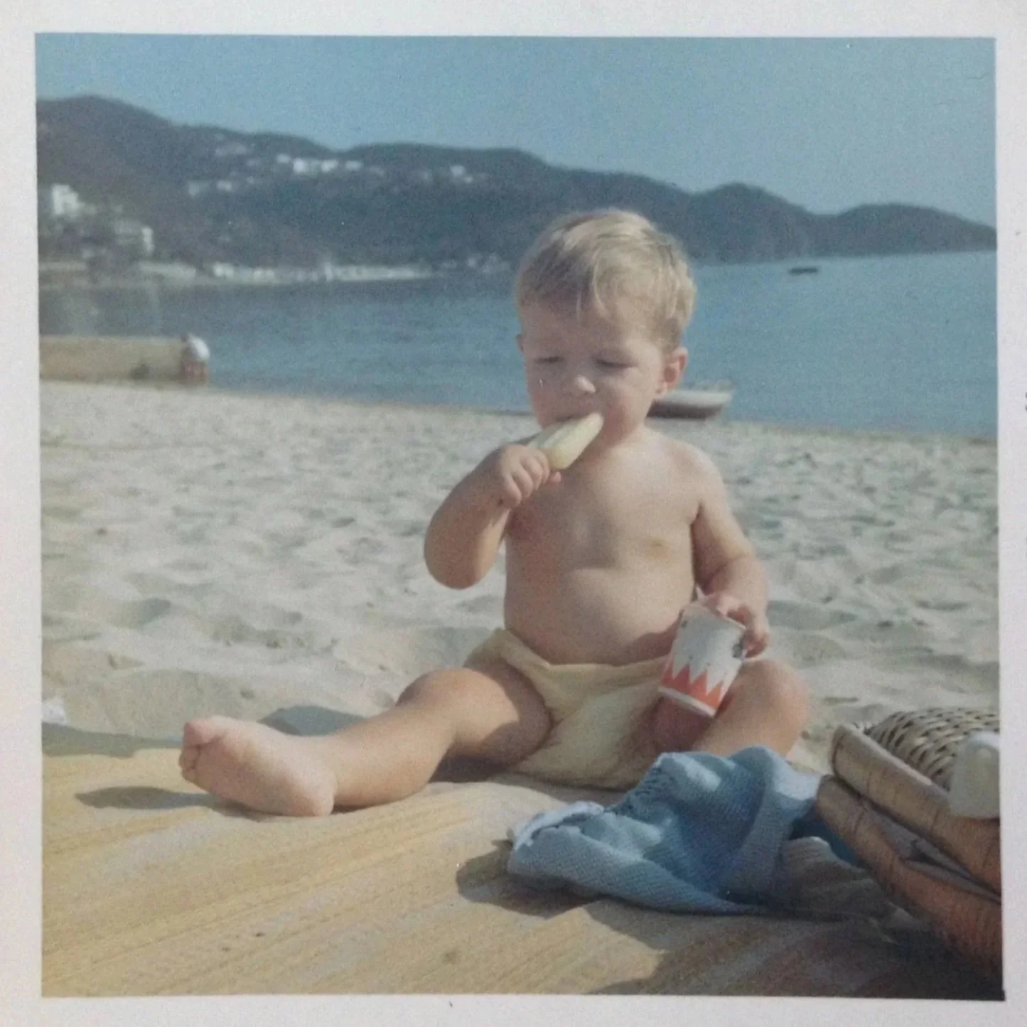 A young boy with light hair sits on a sandy beach eating a banana and holding a paper cup, with a scenic ocean and hills in the background.