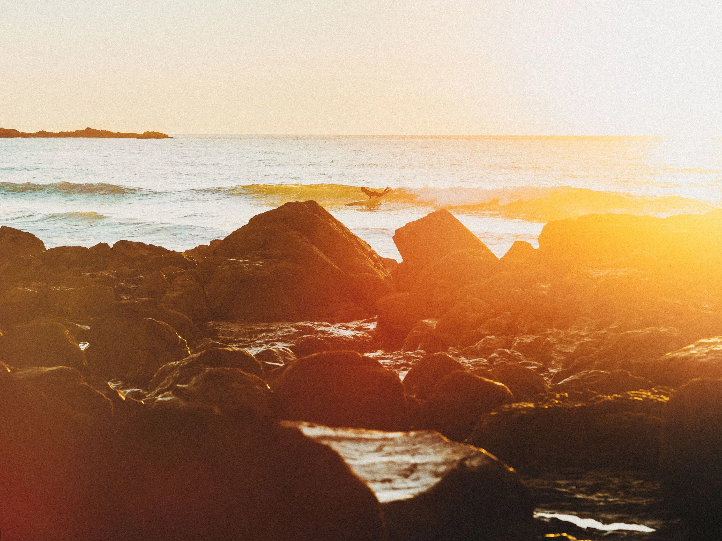 Sunset over the ocean with rocks in the foreground and a surfer in the water.