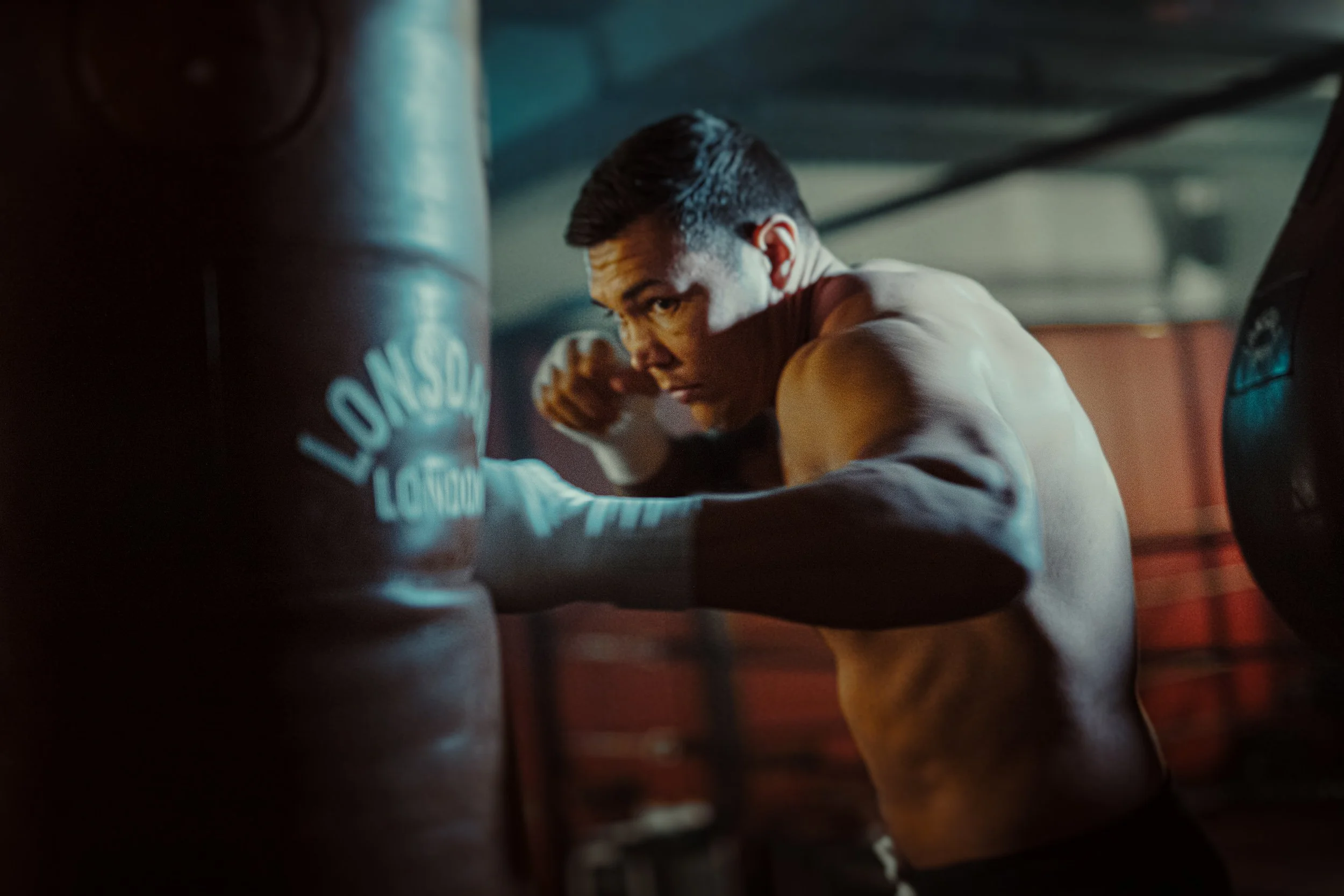 A shirtless man exercising in a dimly lit gym, visible among gym equipment, looking intensely at the camera.