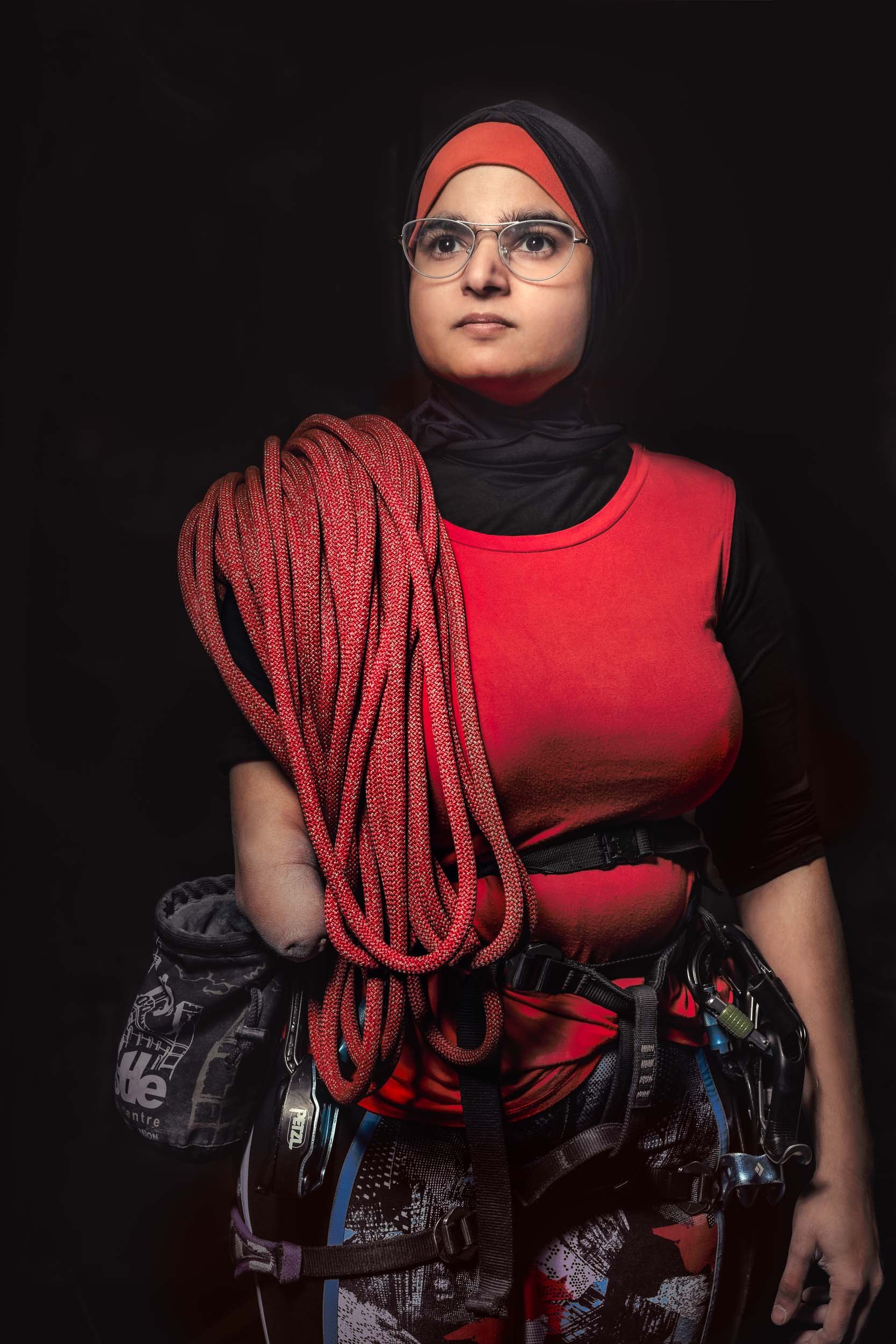 A woman wearing glasses and a hijab with an orange headband, dressed in a red and black climbing outfit, standing against a black background. She is holding a coil of climbing rope over her shoulder and has climbing gear attached to her waist.