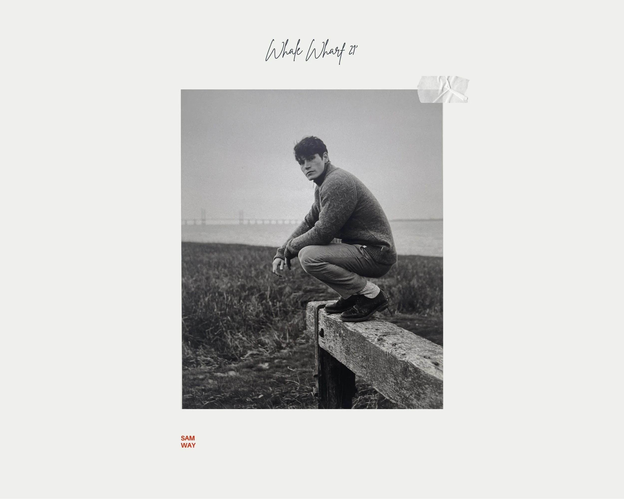 Black-and-white photo of a young man squatting on a wooden beam outdoors near a body of water, with a bridge in the distance, overcast sky, wearing a sweater and pants.