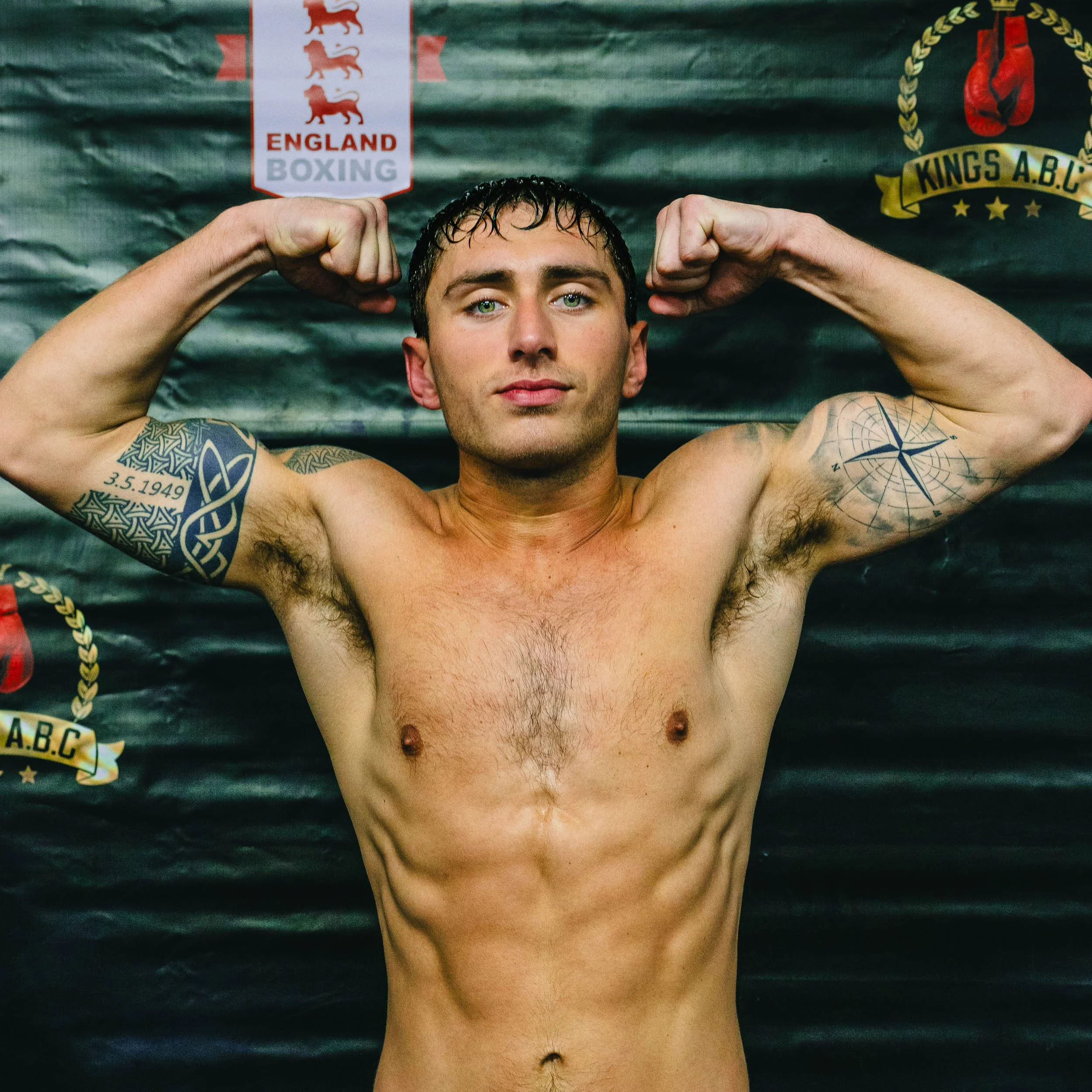 A shirtless man with tattoos flexes his biceps in front of a backdrop with logos of England Boxing and Kings A.B.C, lying on a boxing mat.