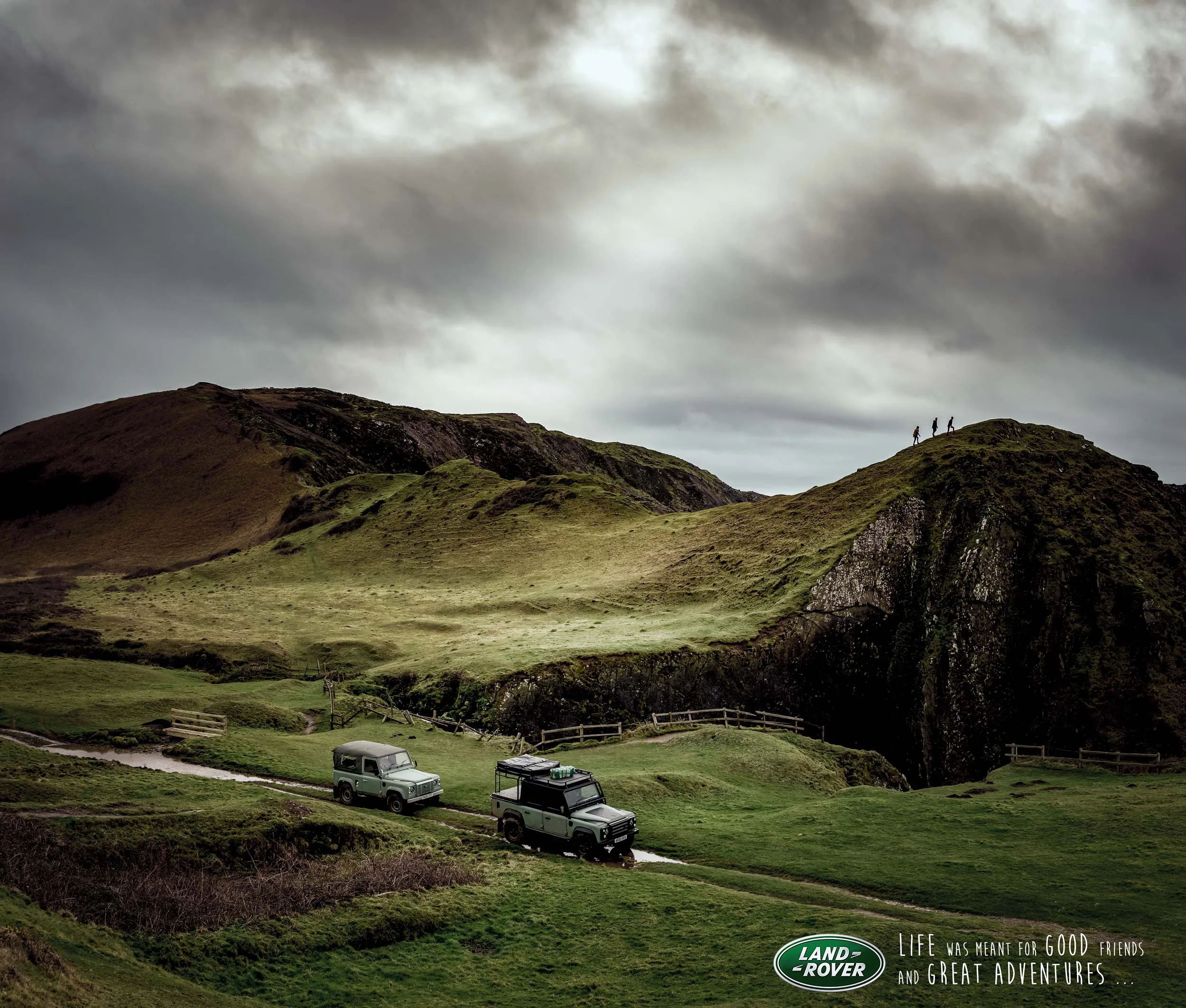 A scenic landscape with green hills, a cloudy sky, and three people hiking in the distance. Two Land Rover vehicles are parked on a grassy area near a small road, along with some wooden fences and a steam of water.