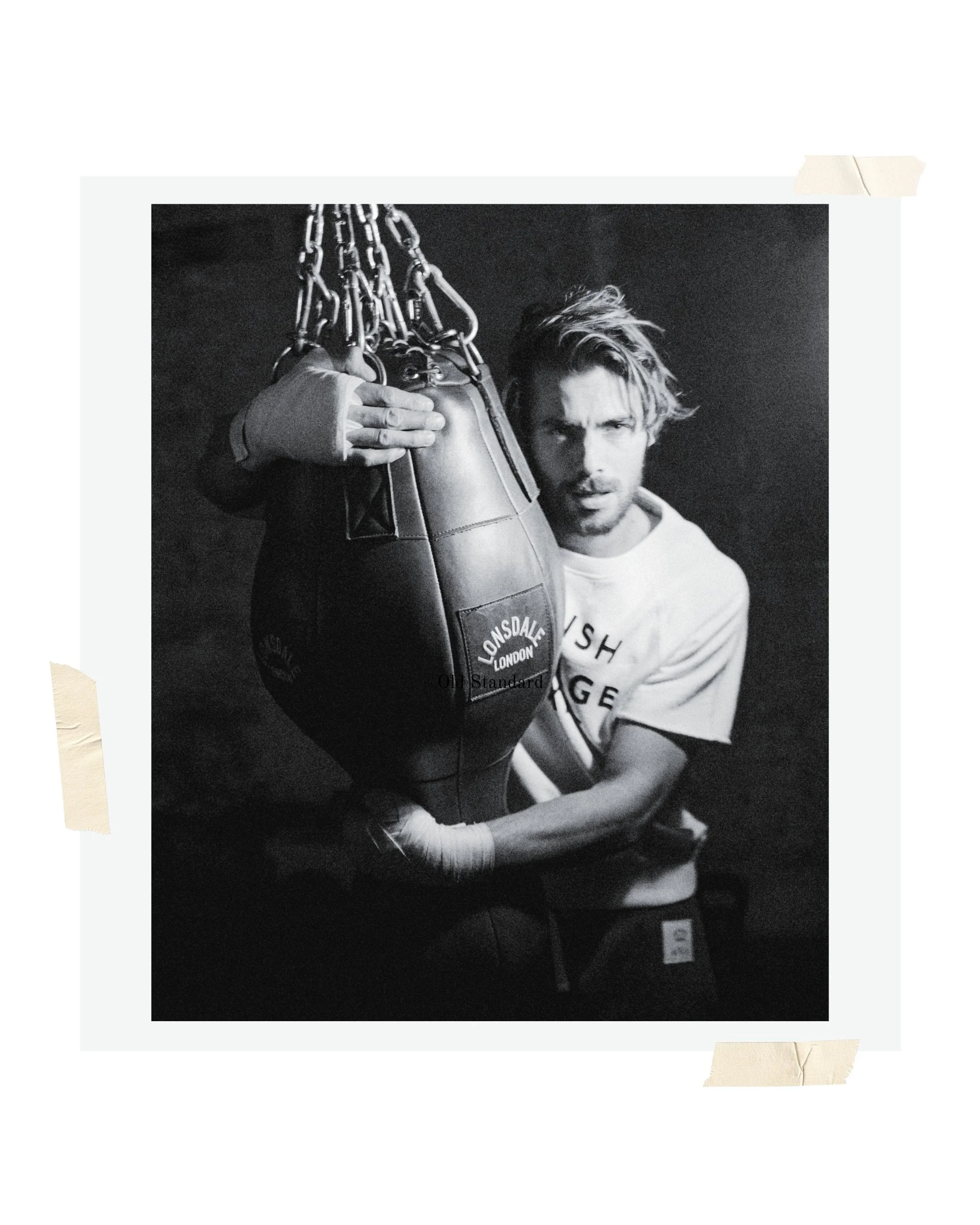 Black and white photo of a man with disheveled hair holding a boxing speed bag labeled 'LONSDALE LONDON,' with a dark background.