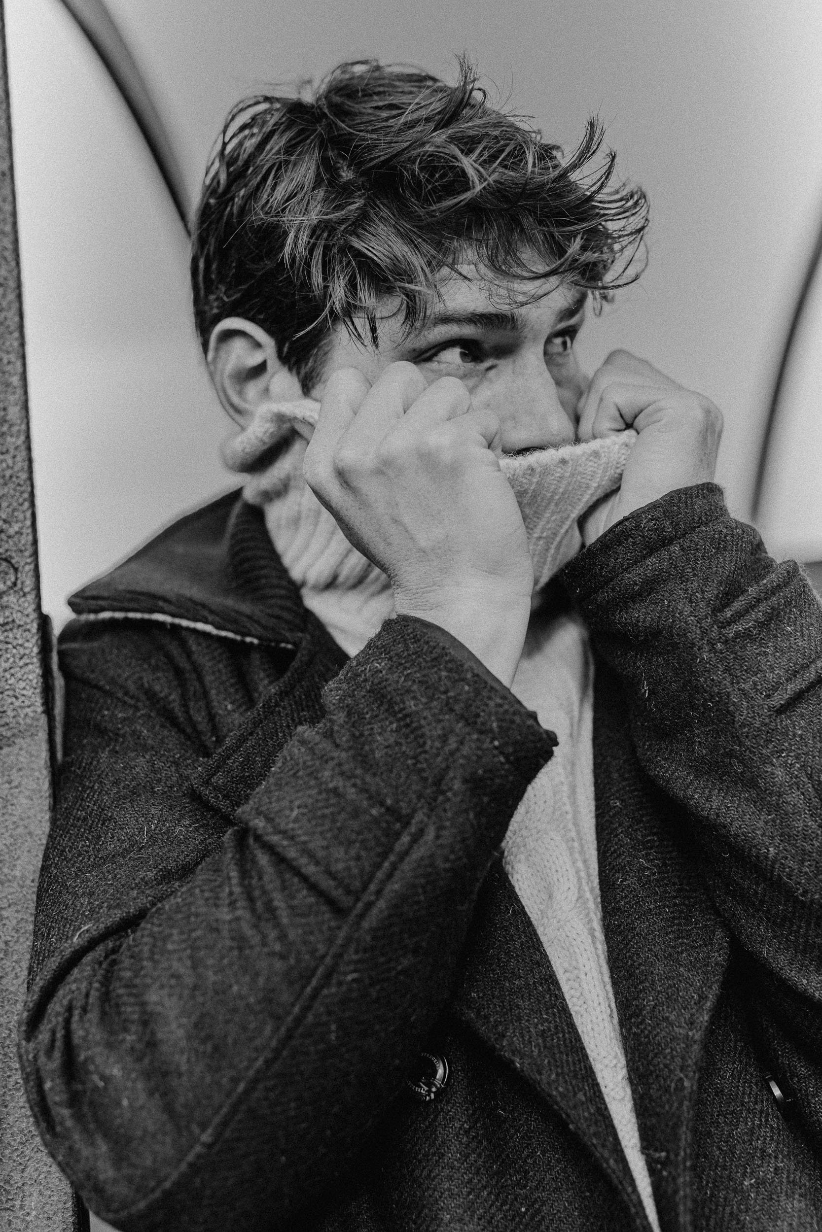 A black and white photo of a person with tousled hair, wearing a dark coat and a sweater, holding a cloth mask over their face while standing against a wall.