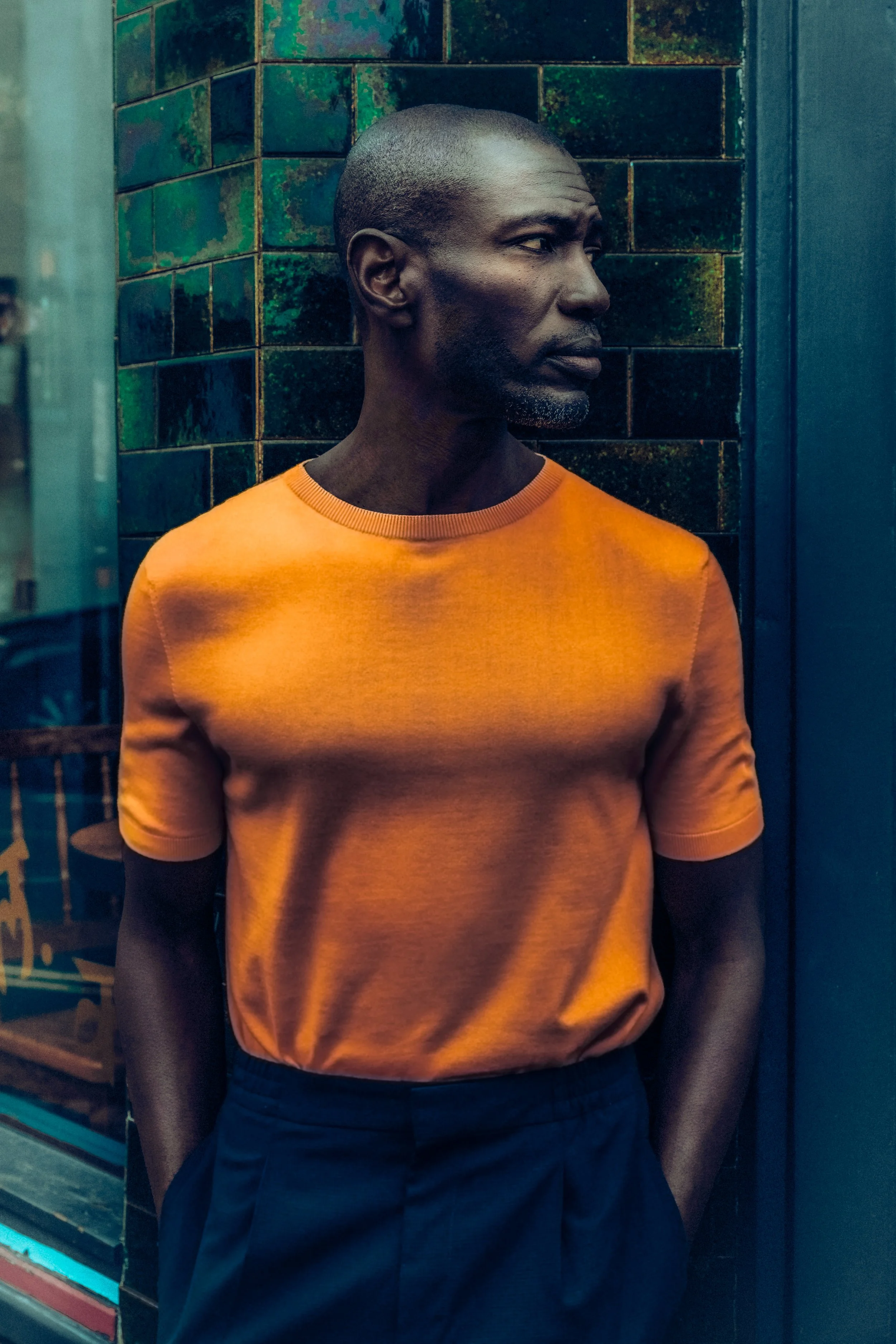 A man with dark skin and a shaved head wearing an orange t-shirt and navy pants standing against a colorful tiled wall background.