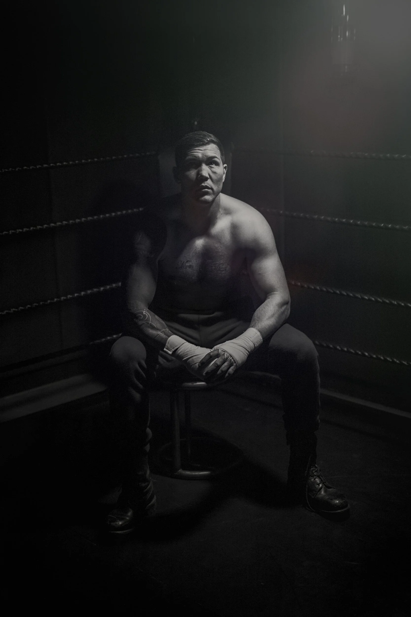 Black-and-white photo of a shirtless man sitting in a boxing ring with black padded walls, looking up with a serious expression.