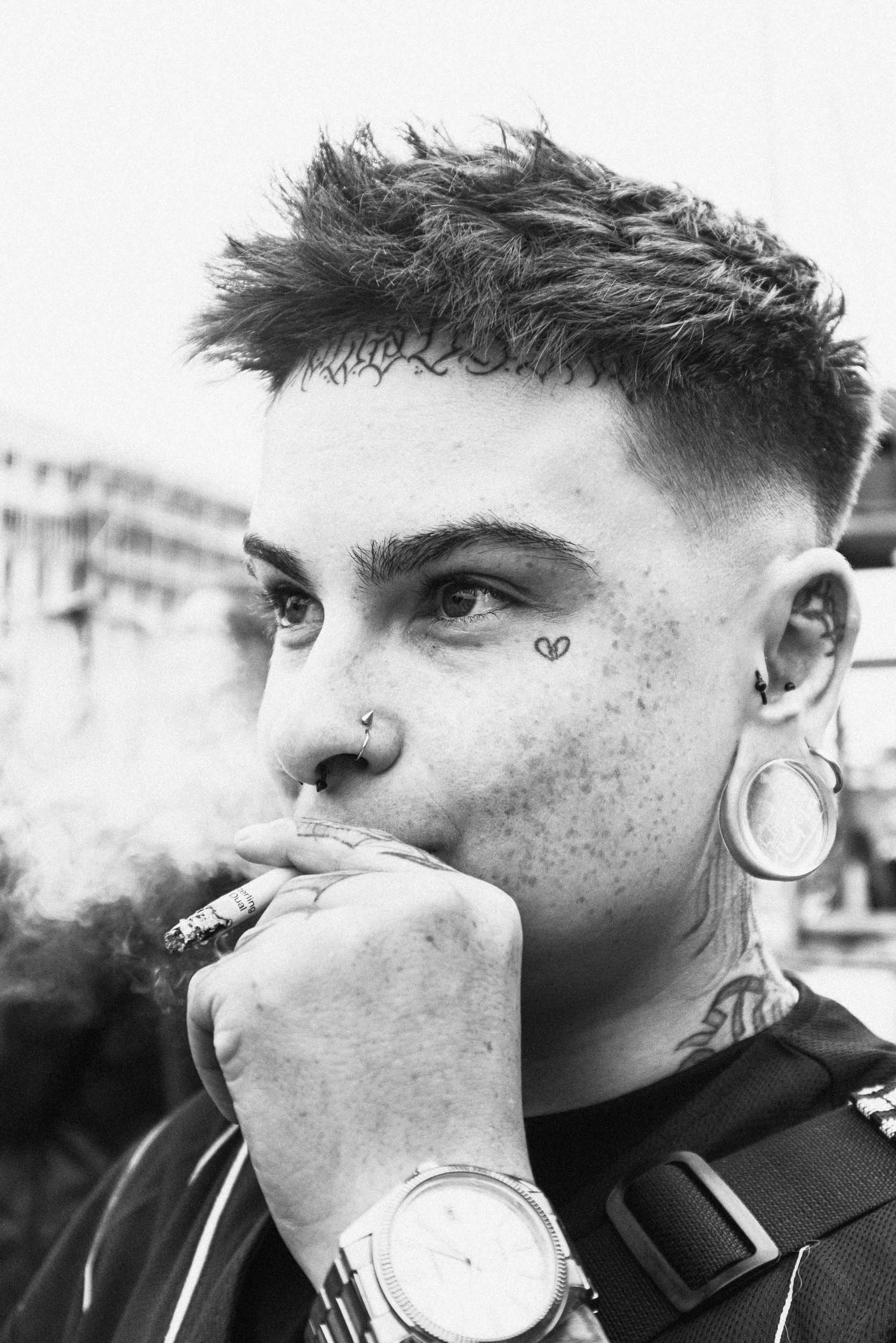 Black and white close-up of a person's face with multiple facial tattoos, including a small heart near the eye and script above the eyebrow. The individual has facial piercings, including a septum ring, and large gauge earrings. They are holding a cigarette near their mouth, wearing a watch and a wristband, with a striped backpack strap visible. The background is blurred with some buildings visible.