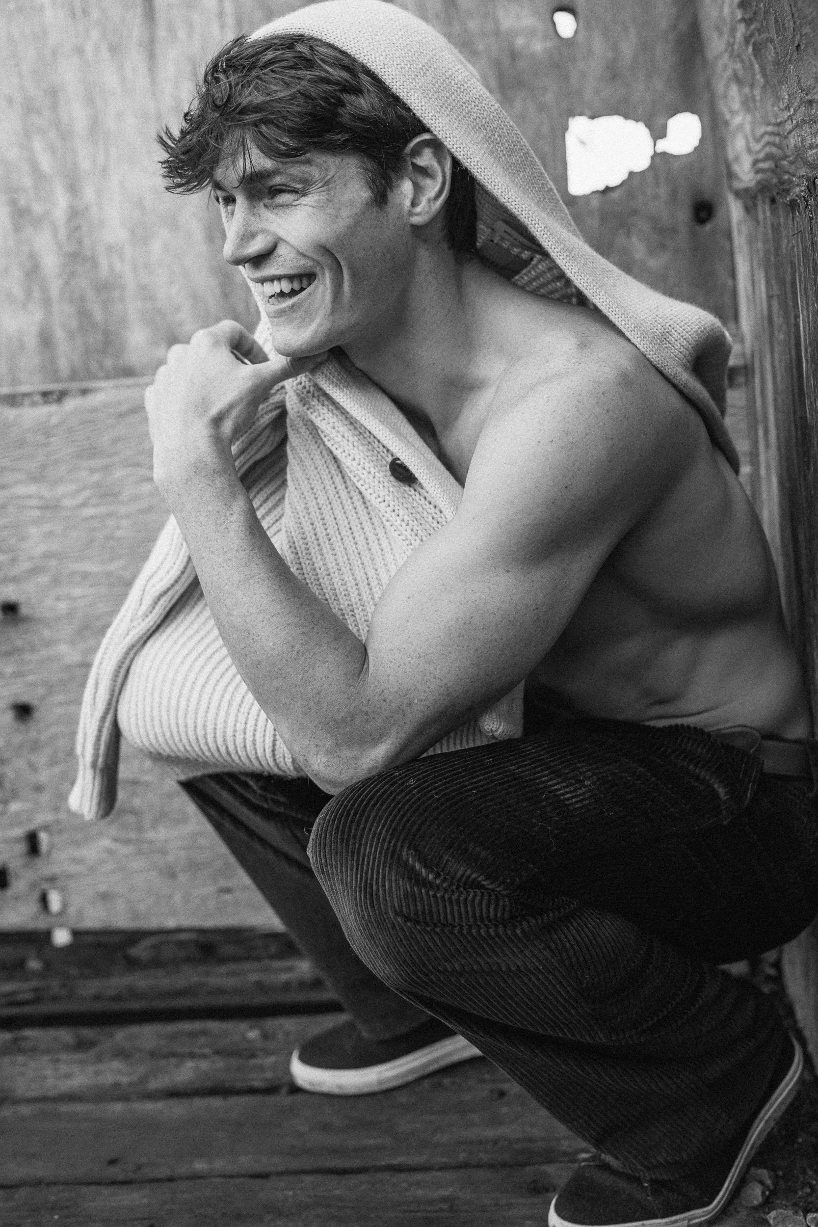 Black and white image of a young man with curly hair, smiling and winking, sitting outdoors near a wooden fence, with a towel draped over his shoulders.