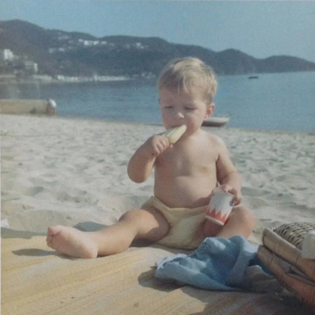 A young child sitting on a sandy beach, eating a banana and holding a container, with boats on the water and a mountain in the background.