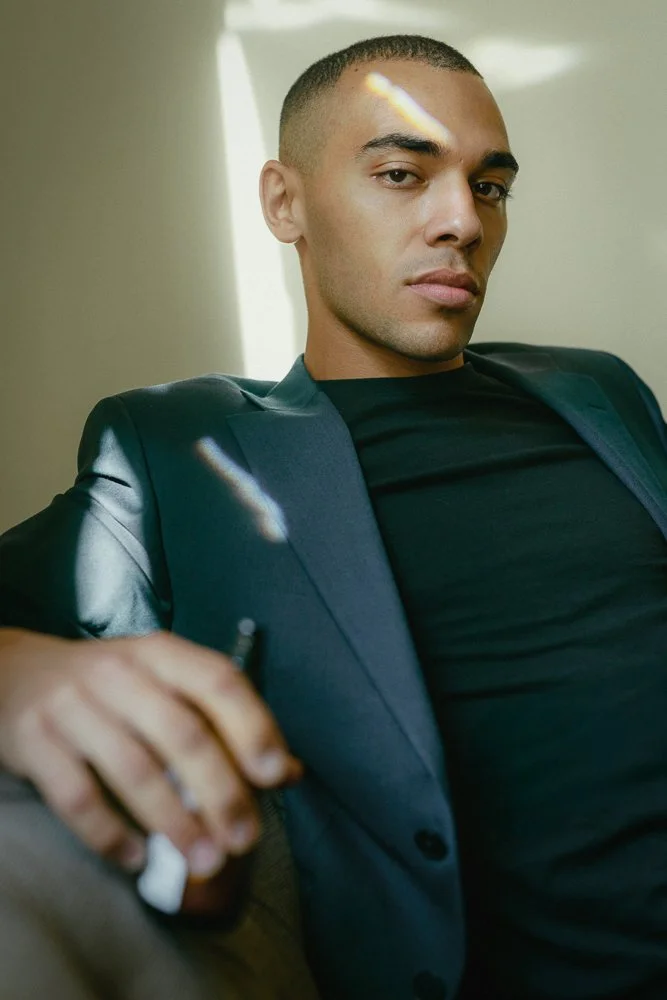 A young man with short hair, wearing a black shirt and a dark blazer, sitting on a beige couch in a room with sunlight. He has a confident expression and is holding a cigarette.