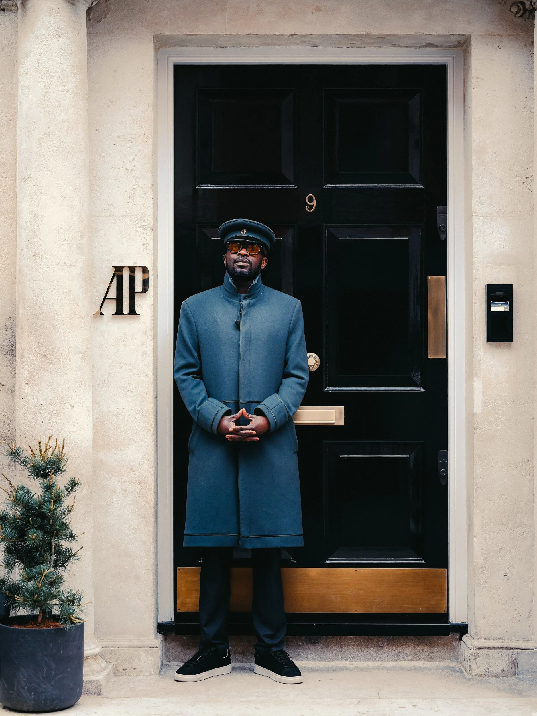 A man dressed in a dark coat, wearing sunglasses and a cap, stands in front of a black door with the number 9, on a building with cream-colored walls. There is a small Christmas tree in a pot to the left.