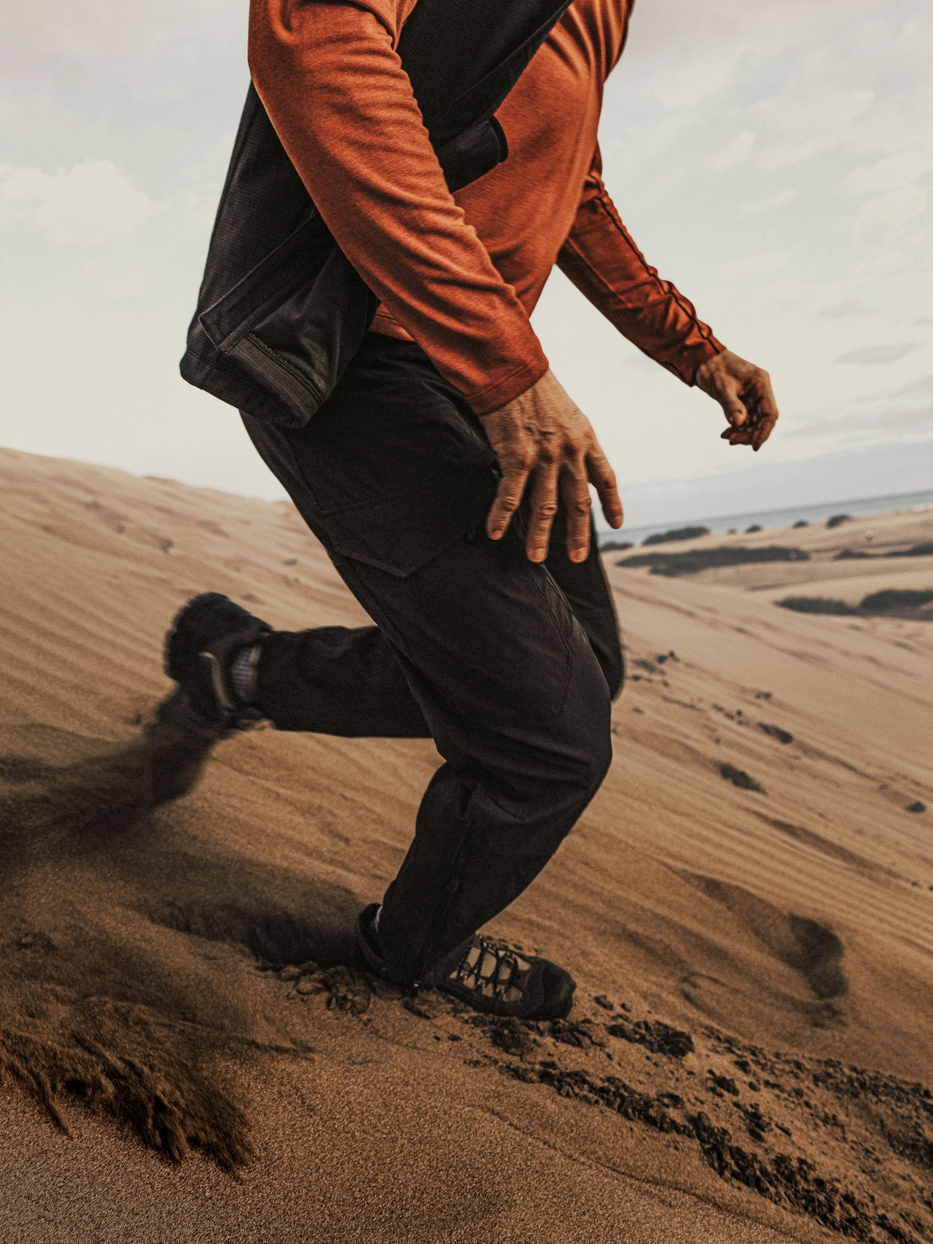 Person wearing hiking boots and outdoor clothing running or walking on sandy terrain with dunes and sky in the background.