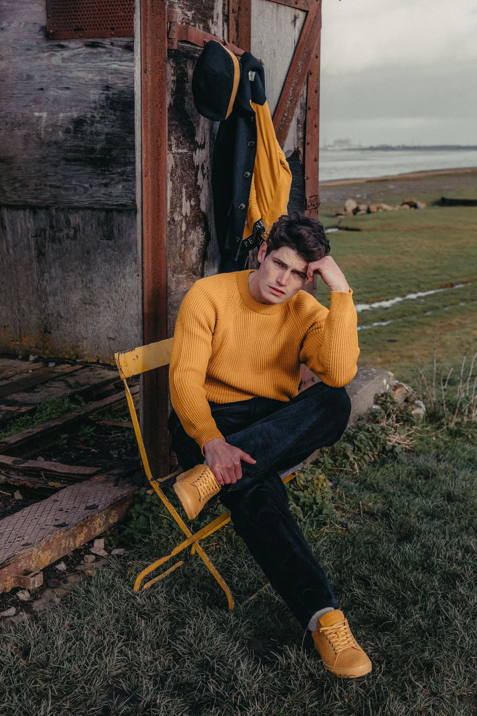 Young man wearing a yellow sweater, yellow shoes, and dark pants sitting on a yellow foldable chair outdoors near a weathered wooden structure, with a background of grass, water, and cloudy sky.