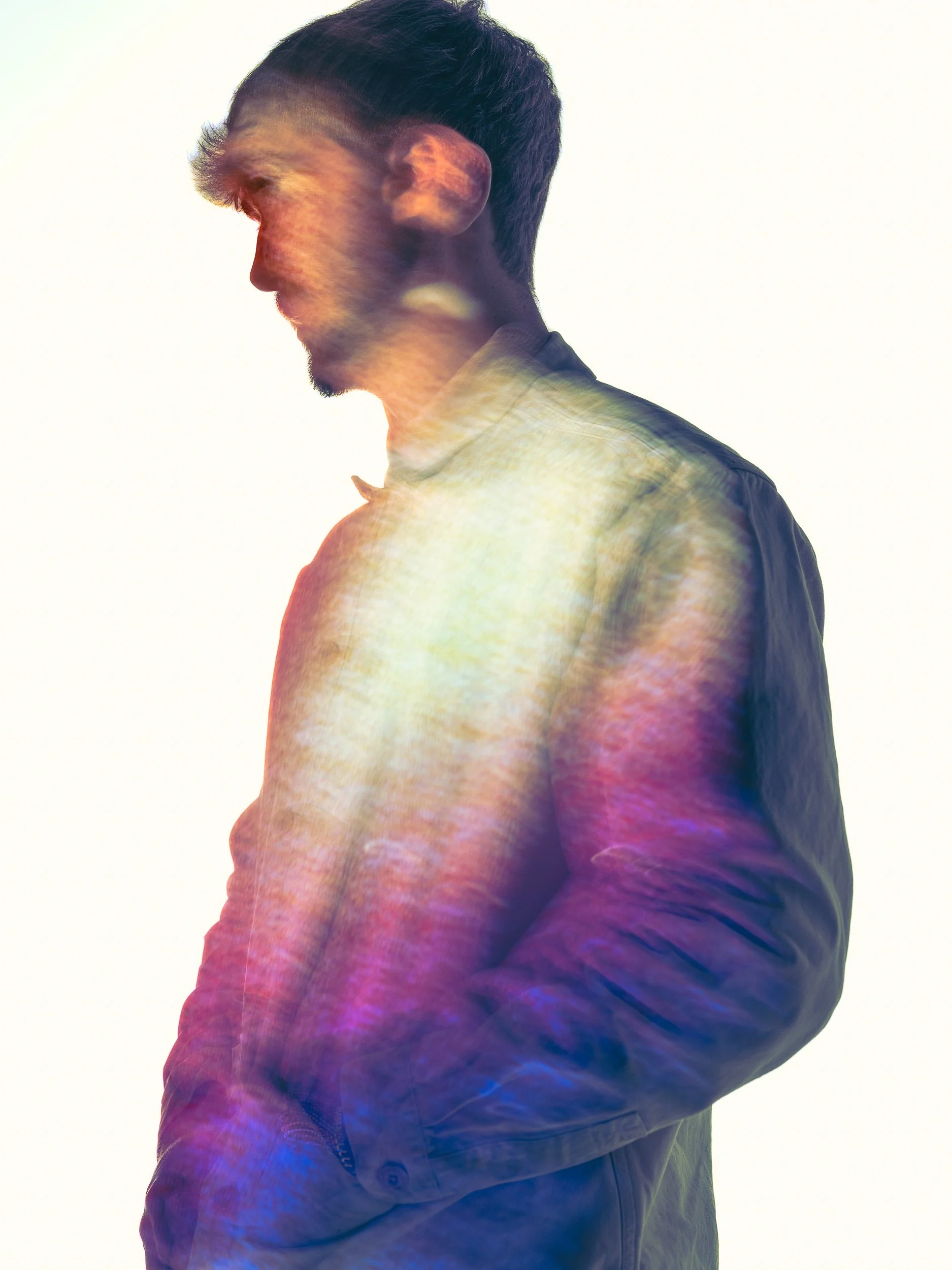 A side profile of a man with dark hair and a beard, wearing a jacket, illuminated by multicolored lights with a white background.