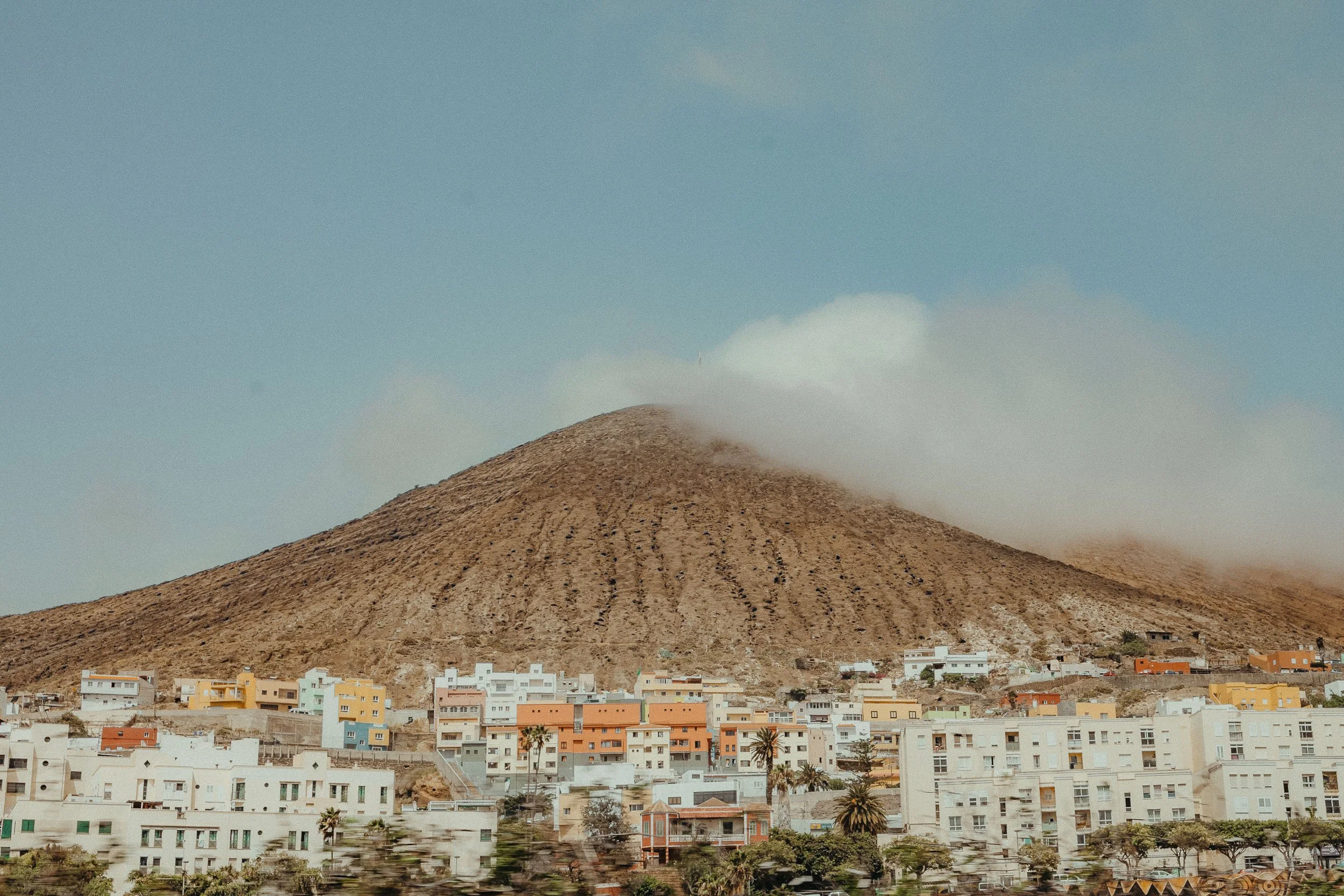 Colorful buildings at the base of a large, brown, volcanic mountain with a cloud at the top in a clear blue sky.