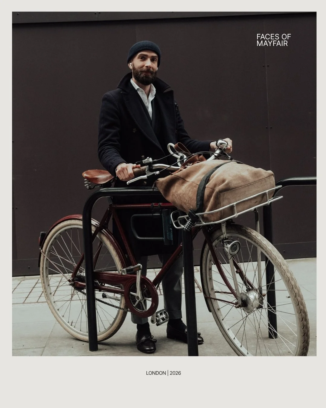 A man with a beard and dark beanie in dark jacket, holding a bicycle with a large brown bag attached to the front, standing in front of a black wall with the text "Faces of Mayfair". The bottom caption reads "London | 2026".