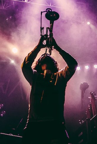 Performer on stage holding a microphone pole, backlit by purple and white stage lights, with smoke and other equipment in the background.