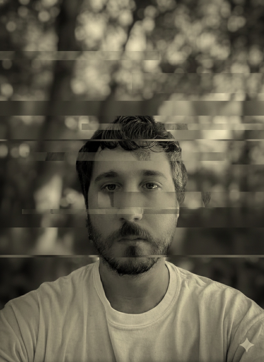 Black and white photo of Andrew Digrius with short hair and a beard, looking directly at the camera, outdoors with blurred trees in the background, with digital glitch effects.