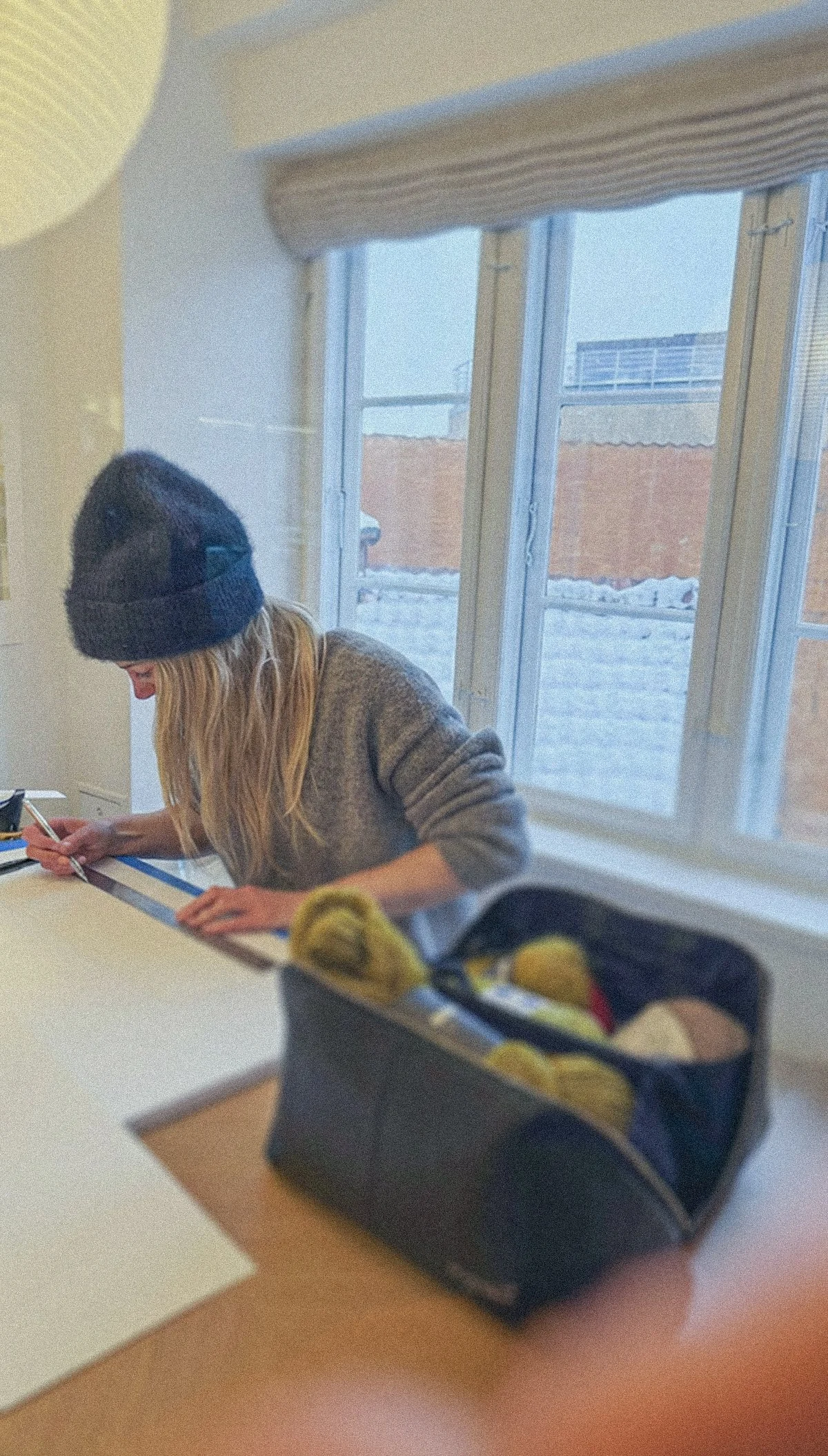 Rebecca Messier sitting at a table in Copenhagen, meticulously measuring stitch lines for a piece of art. A bag with yellow yarn and other art supplies is on the table.