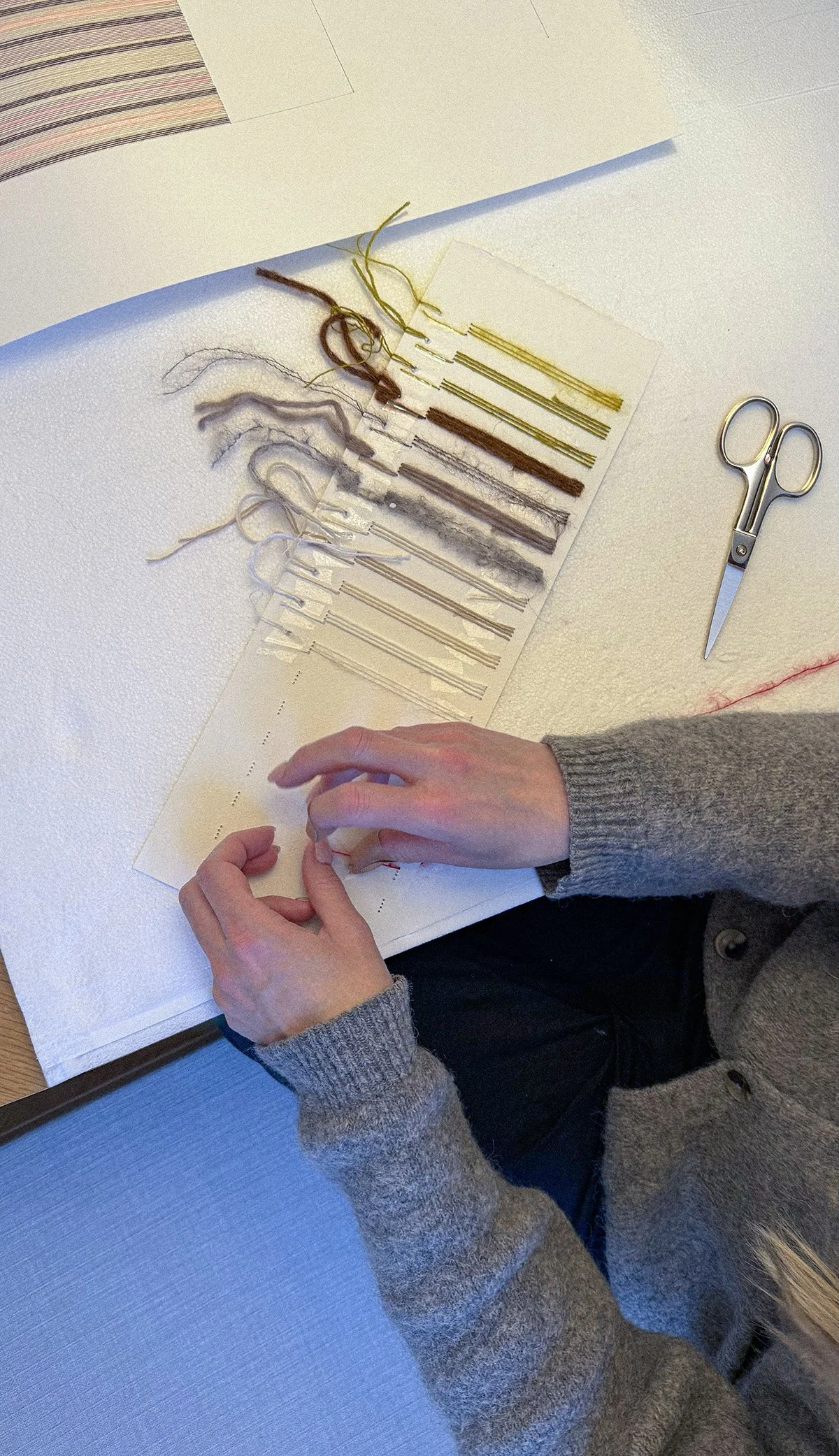 Close up of Rebecca Messier stitching thread and yarn samples onto a paper catalog. Scissors are on the table to the right.