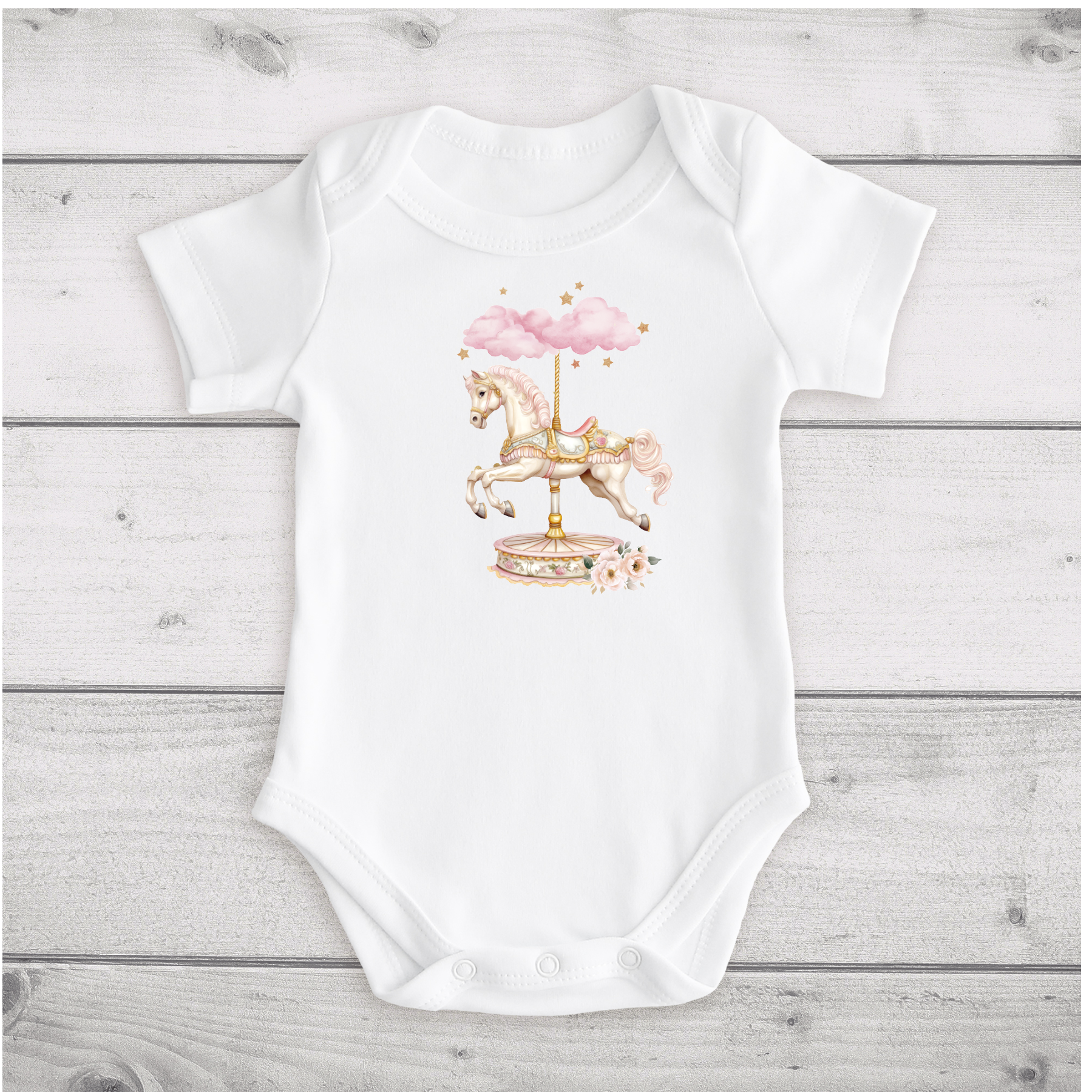 White baby onesie with a colorful carousel horse and pink clouds on the front, lying on a white wooden surface.