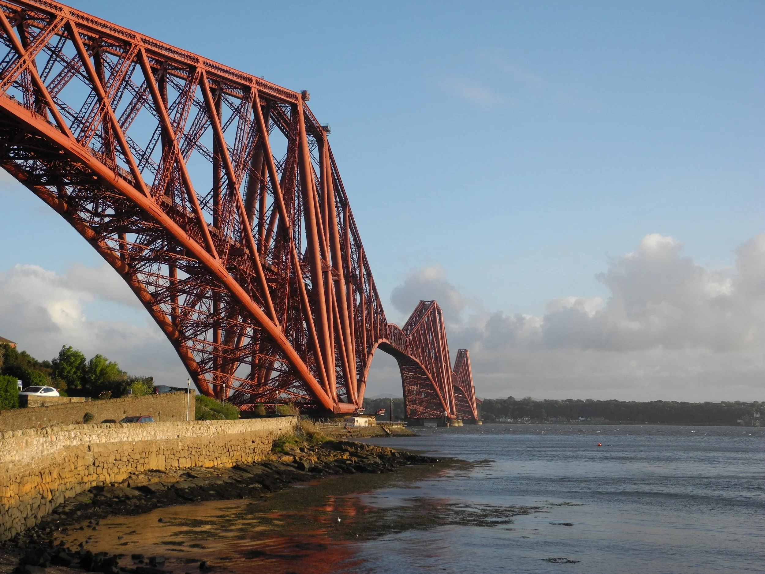 The Forth Bridge, a red steel cantilever railway bridge, spans across the water with a clear sky and some clouds in the background.