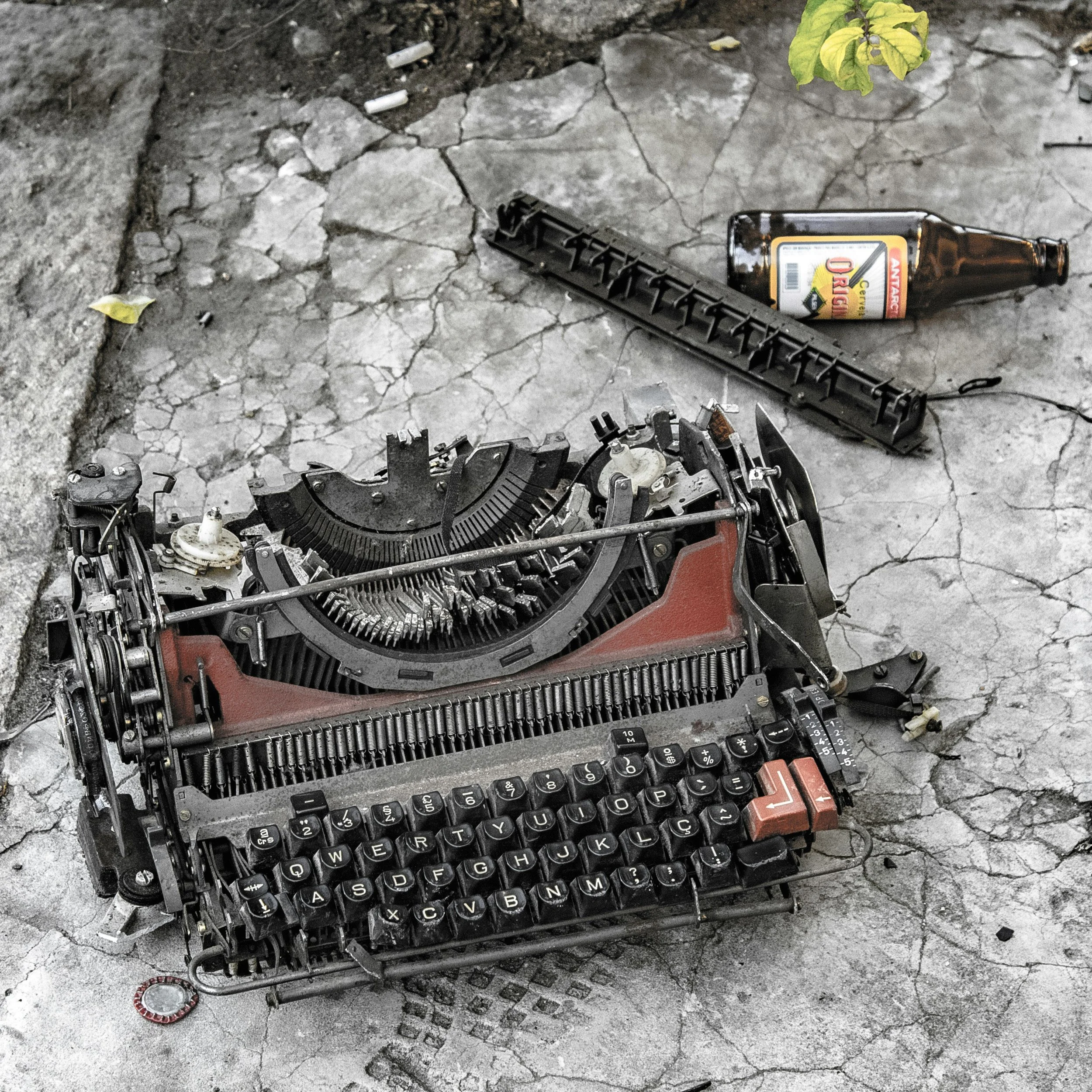A dismantled black typewriter with black keys on a cracked concrete surface. A beer bottle, a black plastic part, and a bottle opener are nearby. There are green leaves in the top right corner.