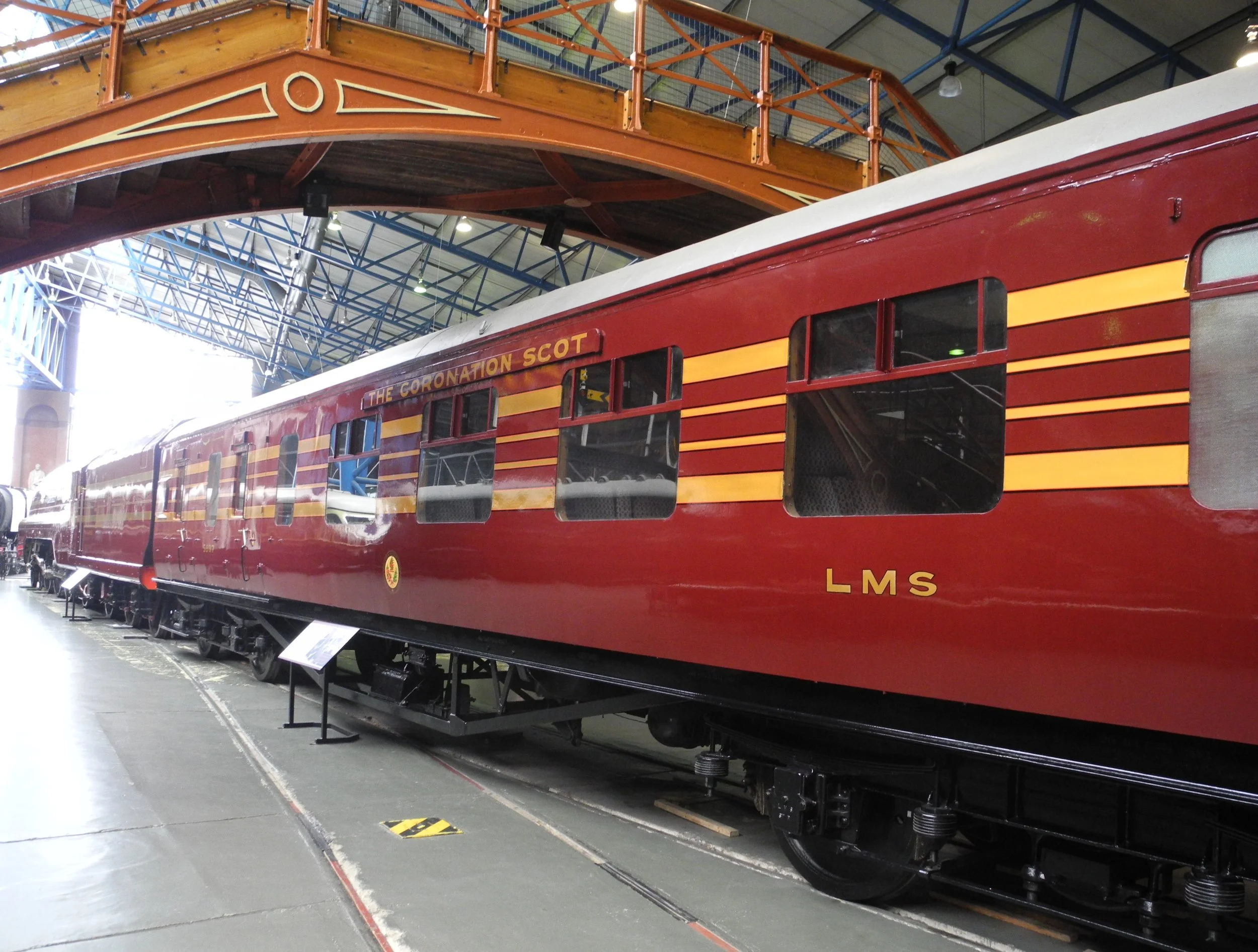 A red train car labeled 'The Coronation Scot LMS' on display inside a museum, with a wooden bridge model and industrial ceiling visible overhead.