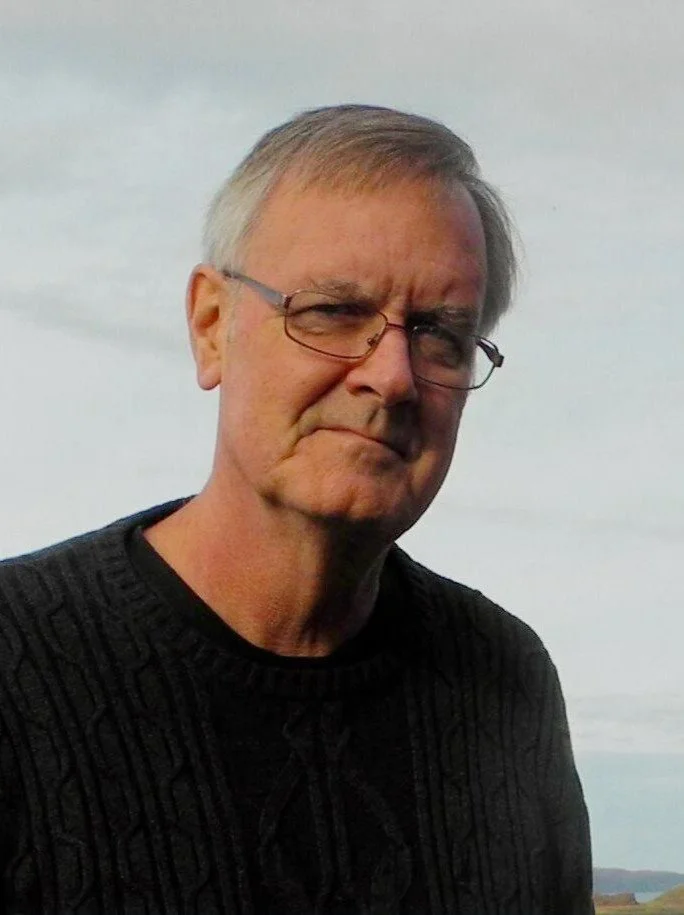 An older man with gray hair, glasses, and a serious expression, wearing a black sweater, outdoors with a cloudy sky in the background.