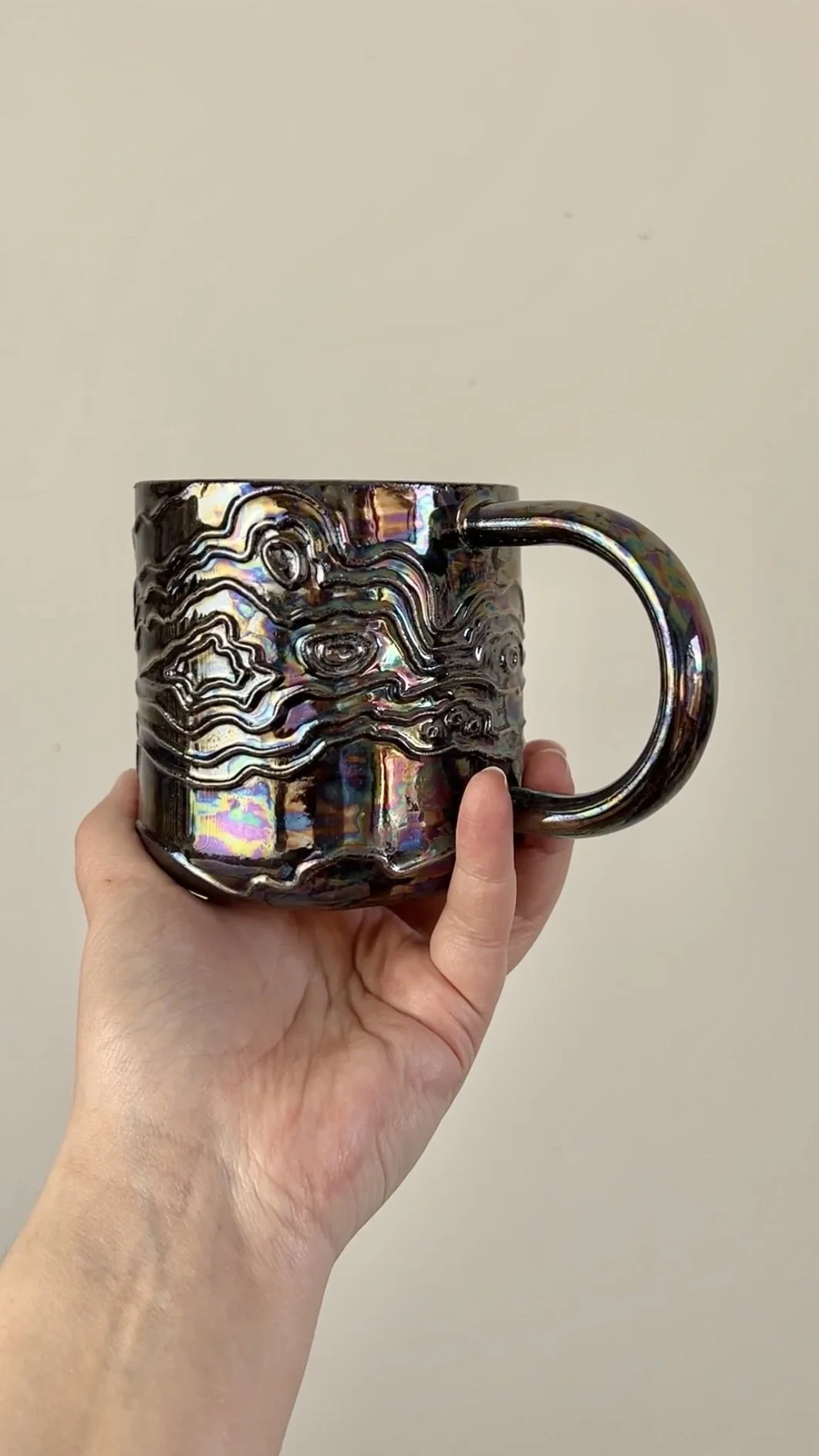 A hand holding a black, metallic, iridescent mug with a textured, topographic pattern against a plain beige background.