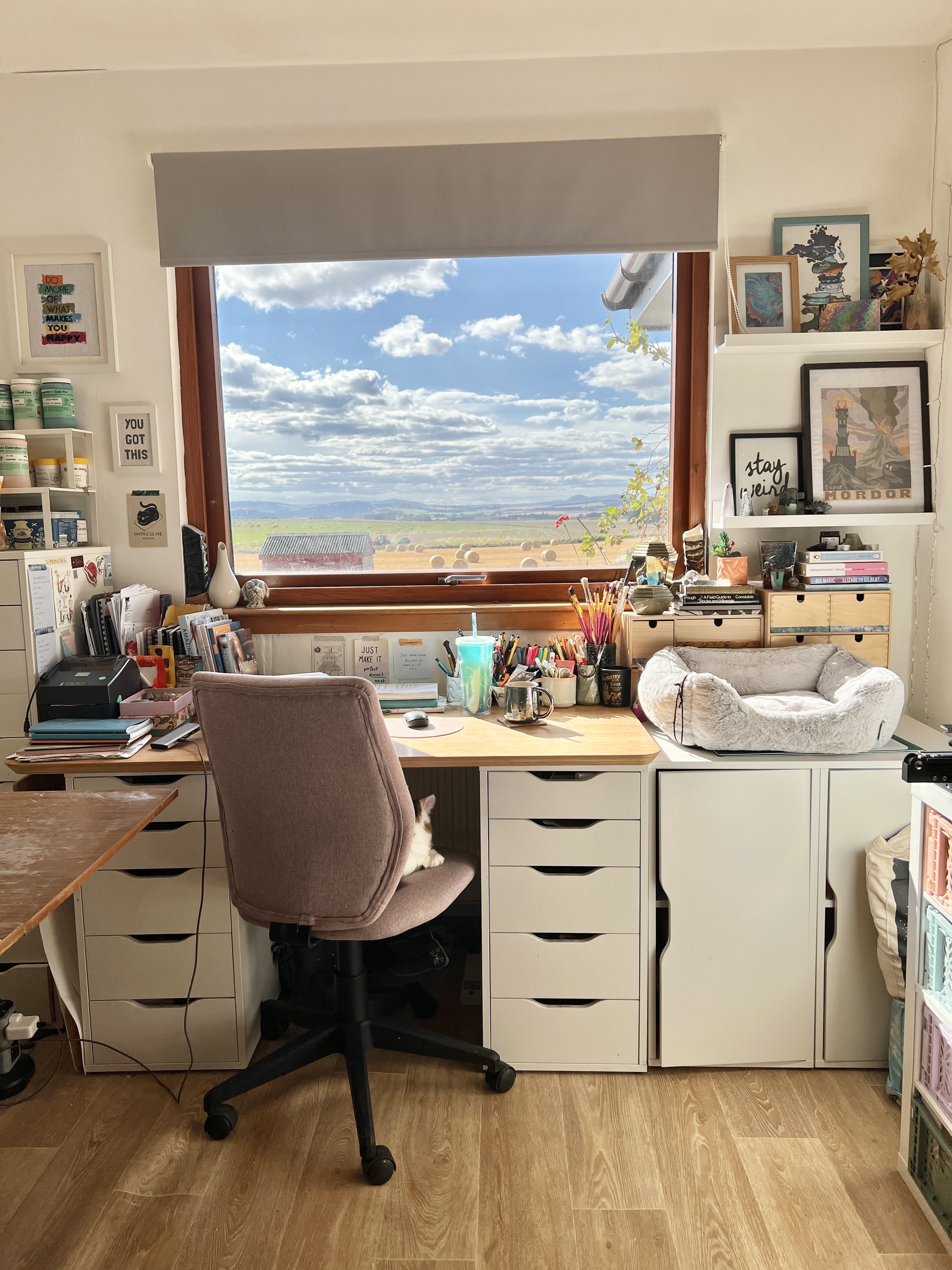 A home office with a desk and an office chair, decorated with art and photos, a window showing a rural landscape with hay bales and blue sky, and various office supplies and plants on the desk and shelves.