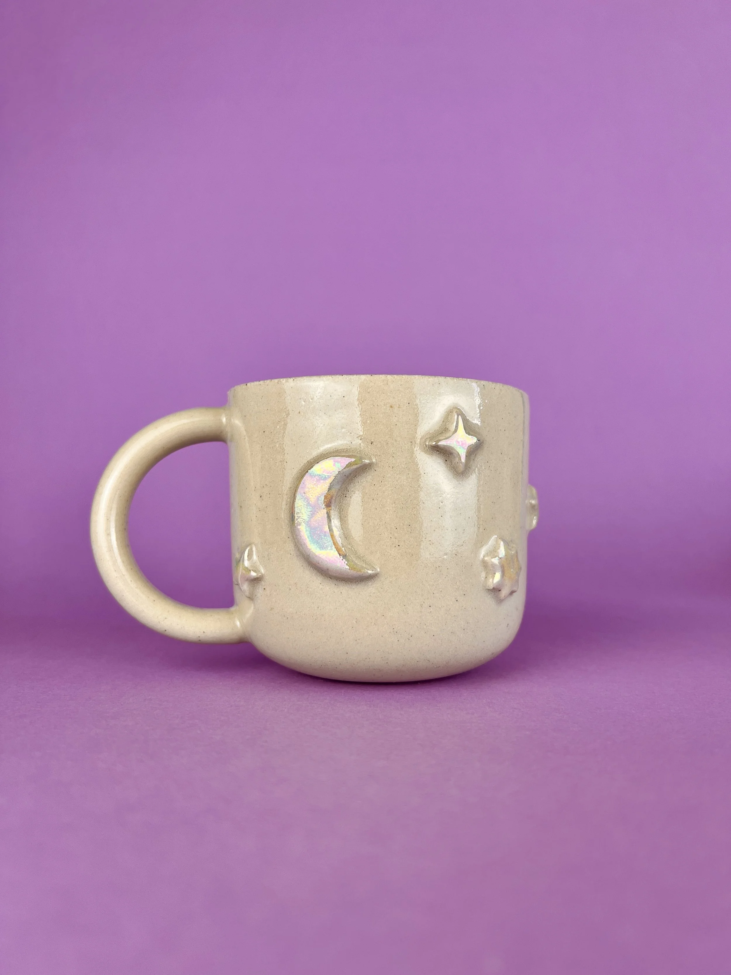 A beige ceramic mug with lunar and star-shaped decorations, set against a purple background.