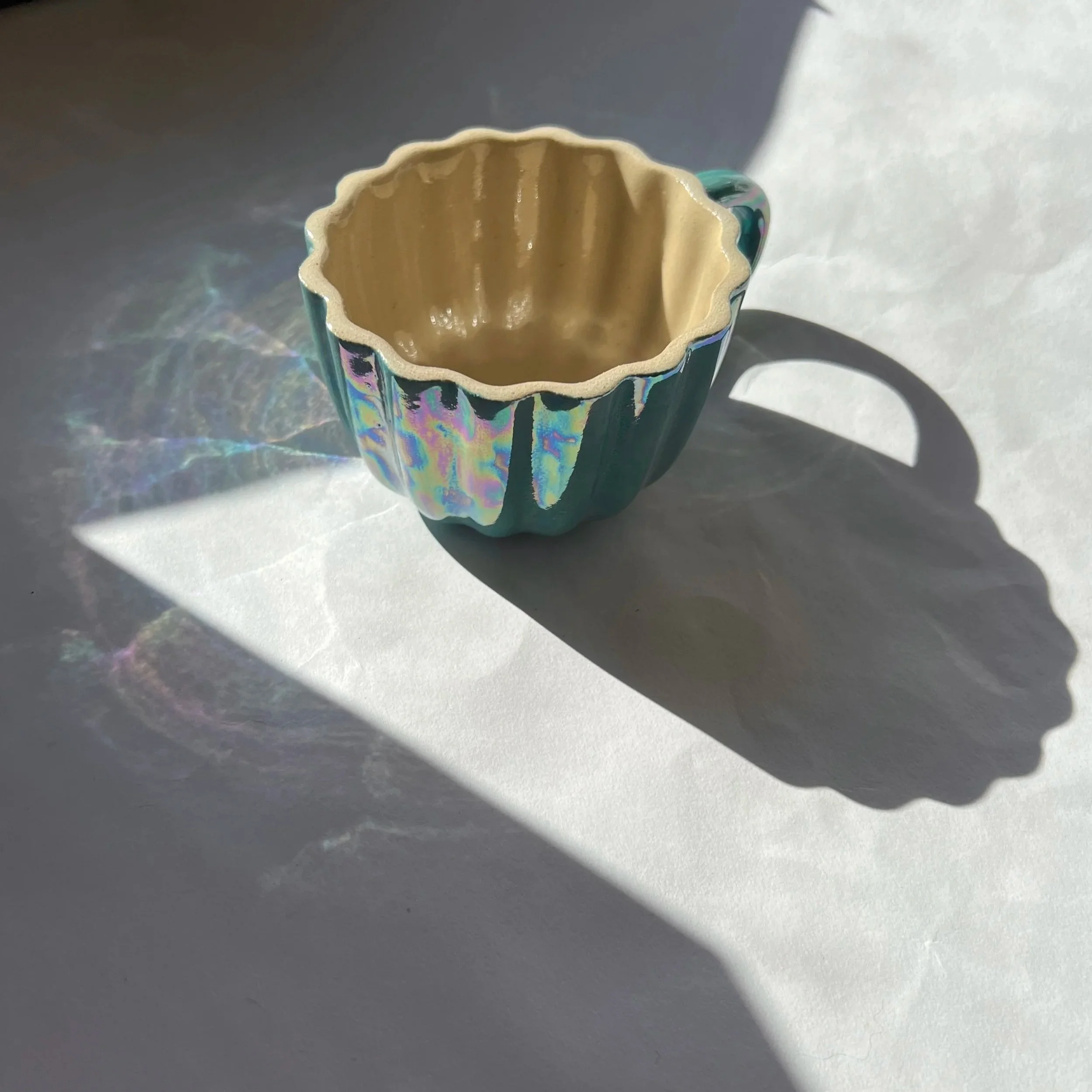 A ceramic cup with an iridescent blue-green glaze on the exterior and a beige interior, casting a shadow and colorful light reflections on a white surface.