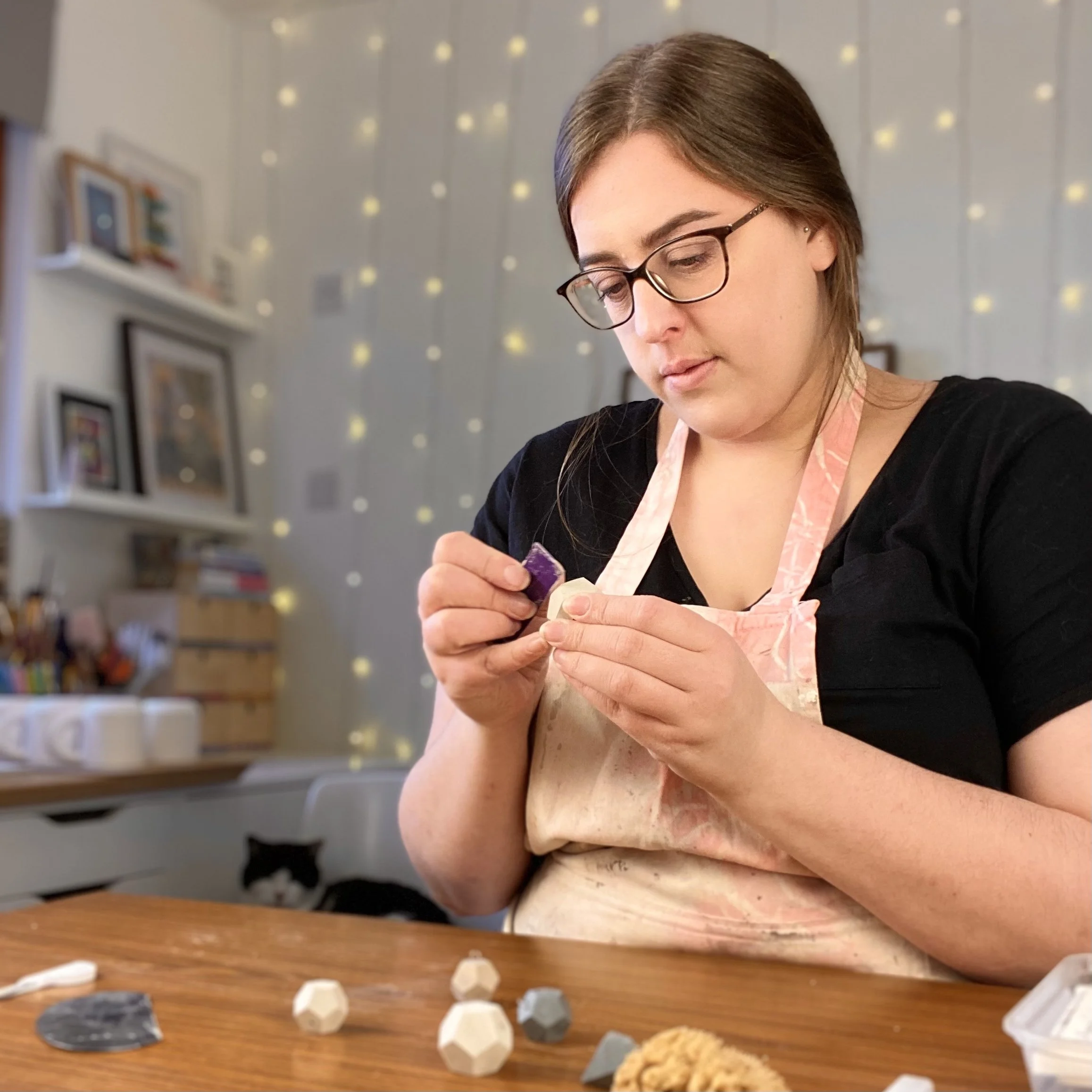 A woman wearing glasses and a pink apron is working on a craft project at a wooden table. There are various craft materials and soap bases on the table, and a black and white cat is resting in the background.