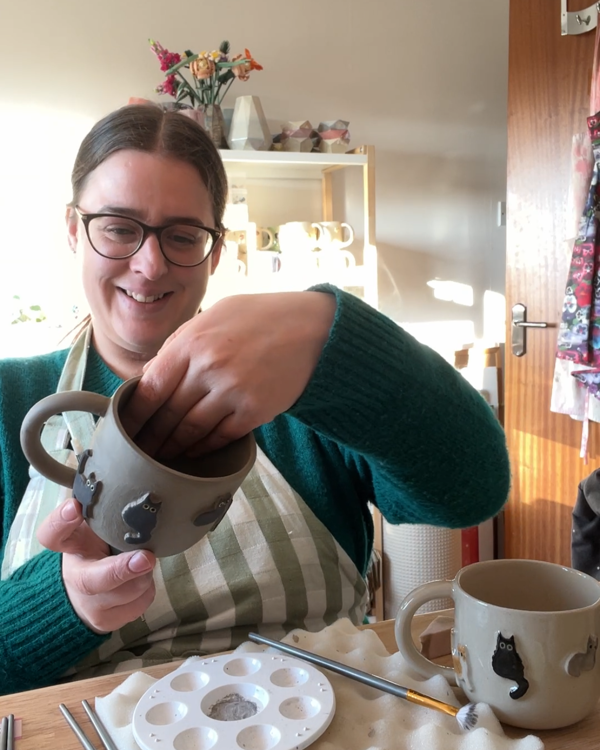 A smiling woman in glasses decorates ceramic mugs with cat designs at a table, with art supplies and finished mugs visible.
